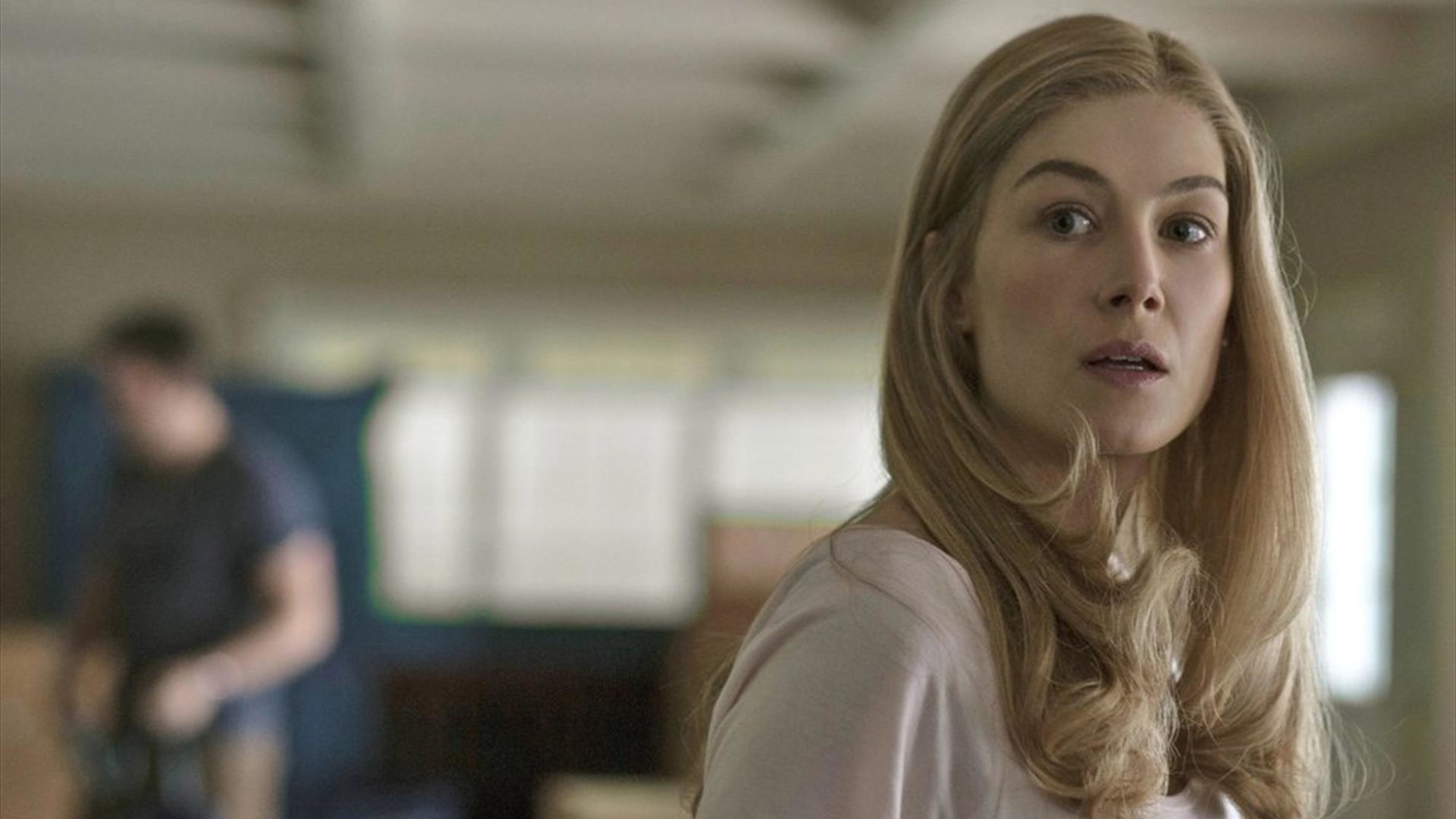 Rosamund Pike with long hair, wearing a light top, looks to the side with a background showing a dimly-lit room