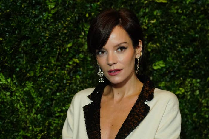 Lily Allen stands against a leafy backdrop, wearing a white outfit with a black collar and drop earrings