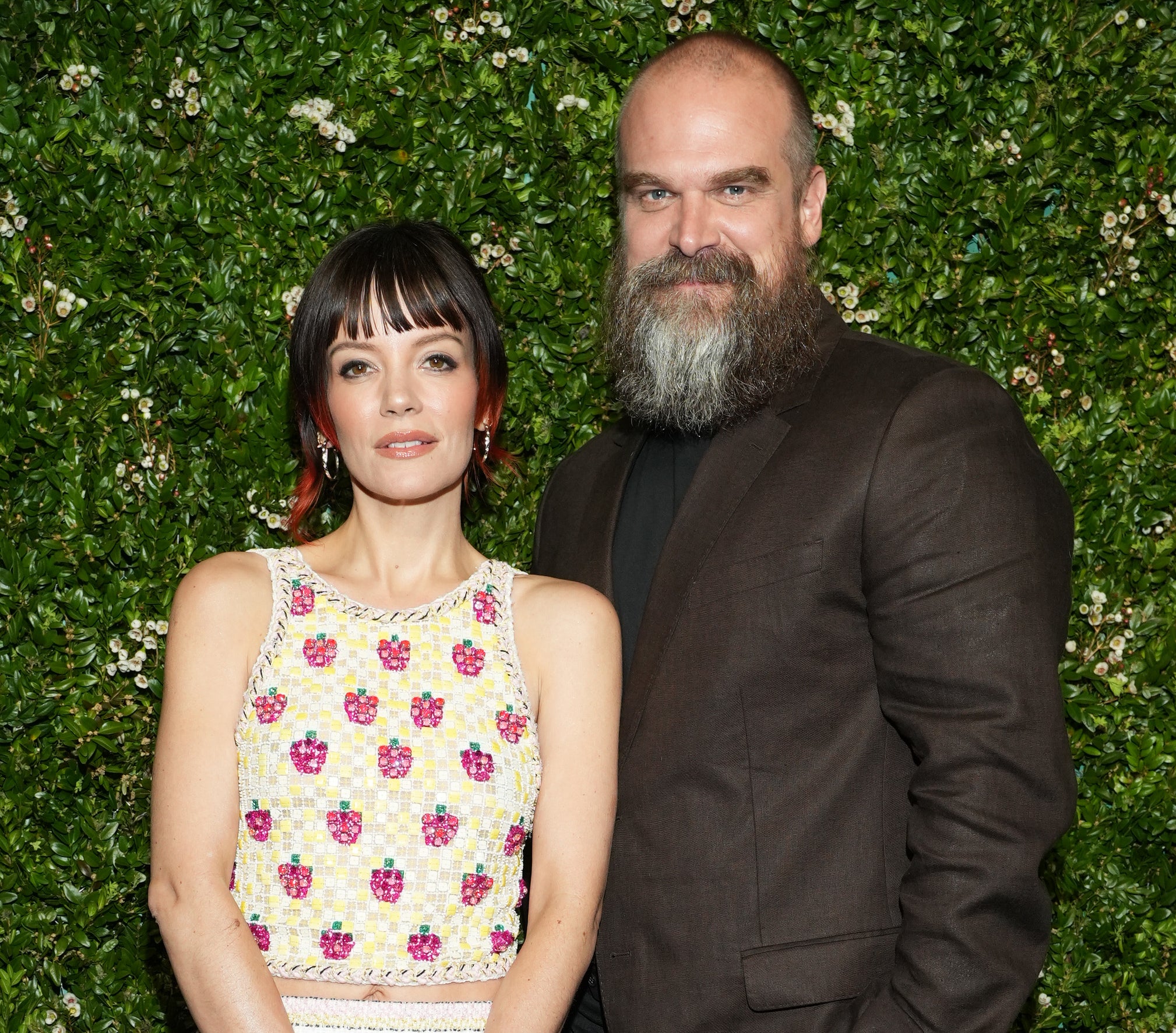 Lily Allen in a floral top and striped skirt, and David Harbour in a dark suit, pose together in front of a leafy backdrop