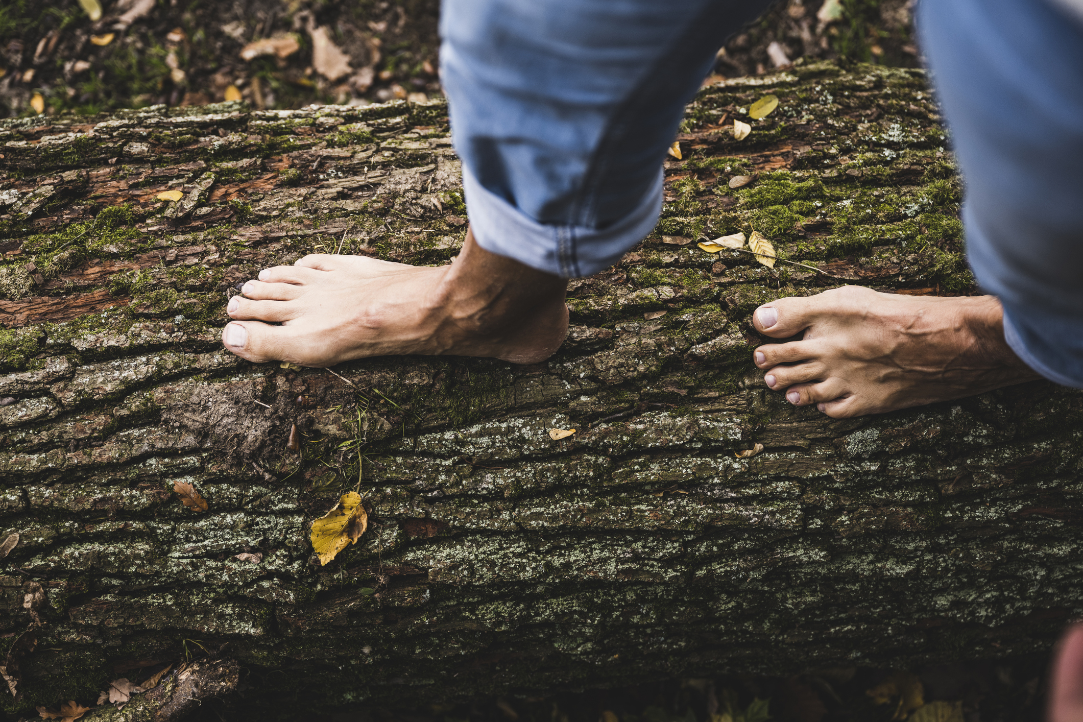 Bare feet walking on a mossy tree log outdoors, with blue rolled-up jeans visible