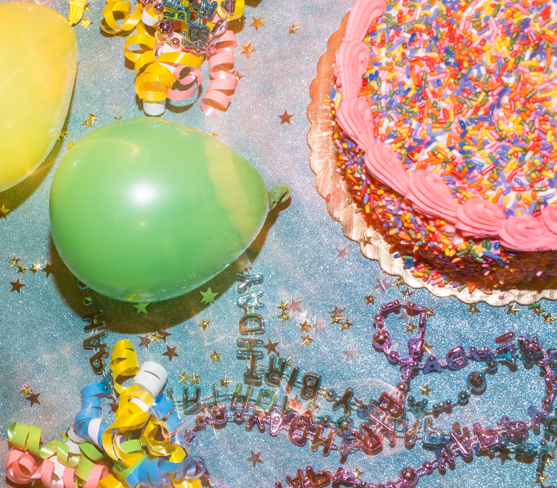 A festive birthday party setting with a cake covered in sprinkles, yellow and green balloons, colorful streamers, and small star confetti