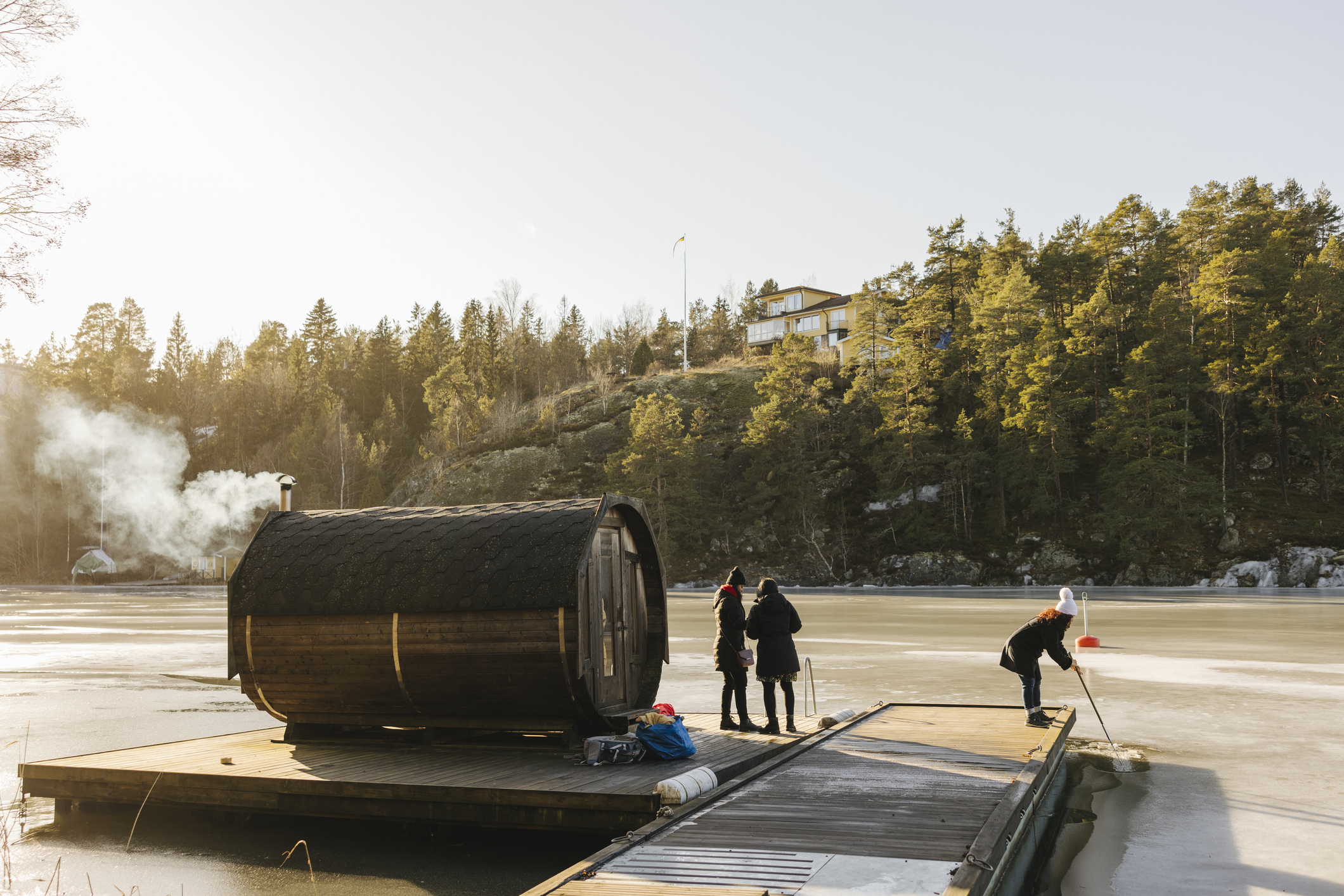People stand on a dock next to a small round sauna cabin by a lake, surrounded by a forested landscape