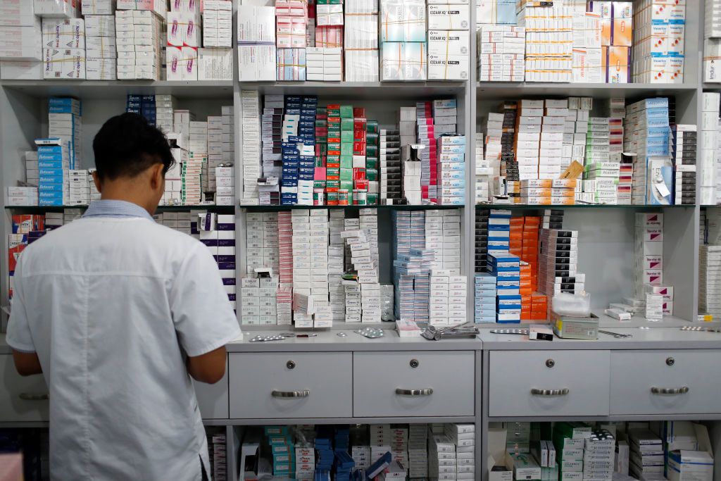 A person wearing a white coat is standing in front of shelves filled with various medications in a pharmacy