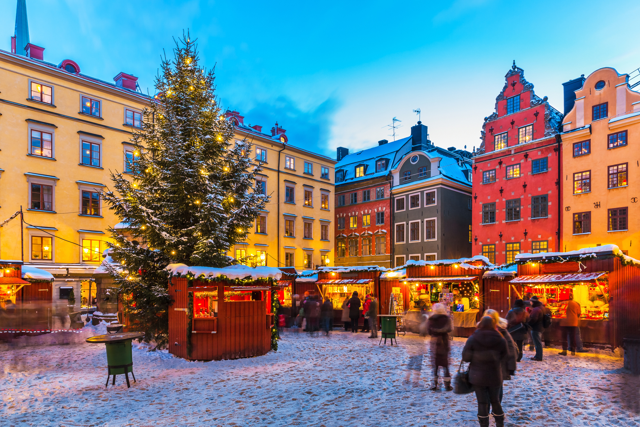A lively Christmas market in a snowy town square with various vendor stalls, a decorated large Christmas tree, and colorful historic buildings in the background