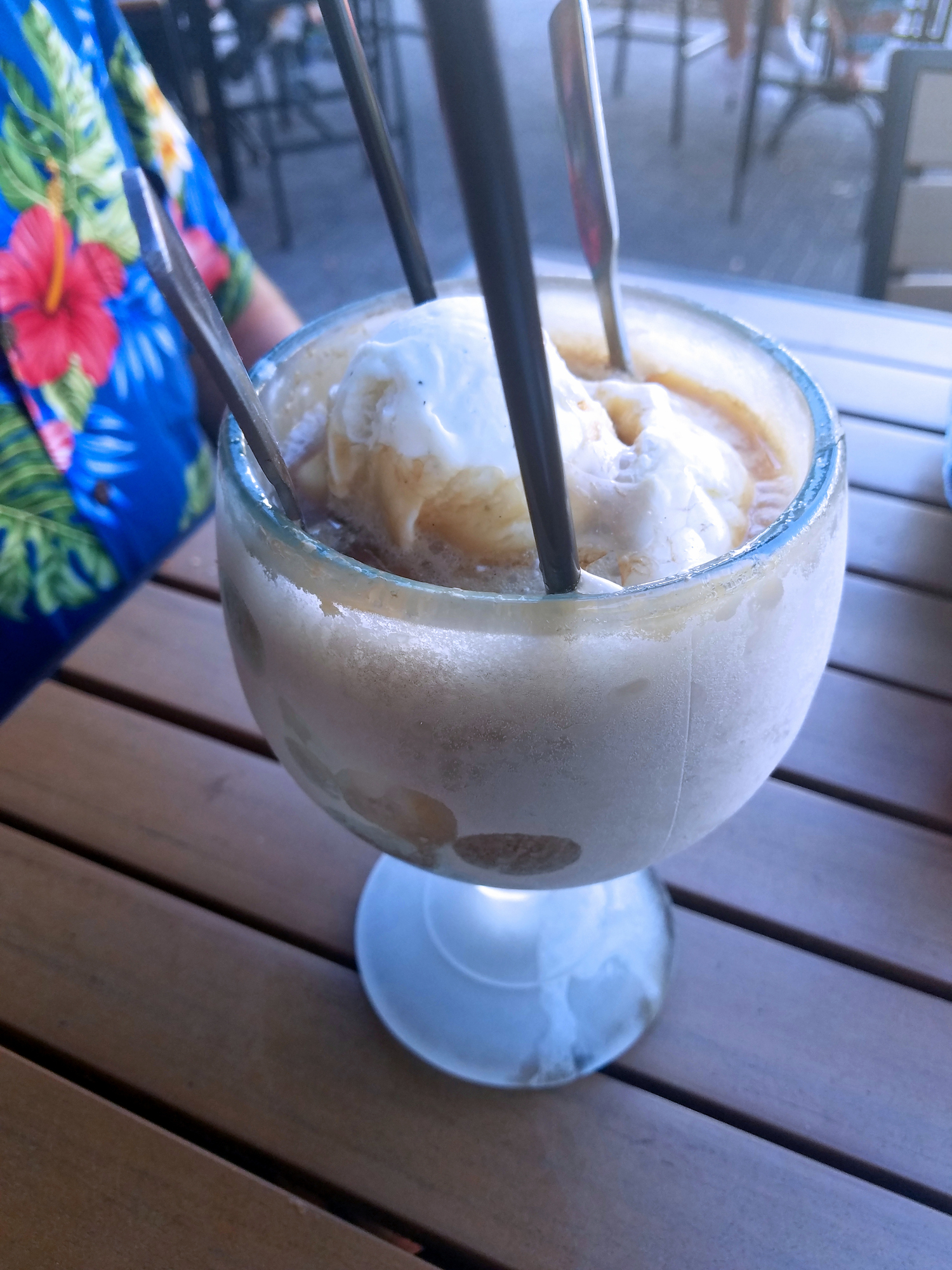 Large frosted glass filled with ice cream and a drink, topped with whipped cream. Three straws and two spoons are in the glass. Person in floral shirt nearby