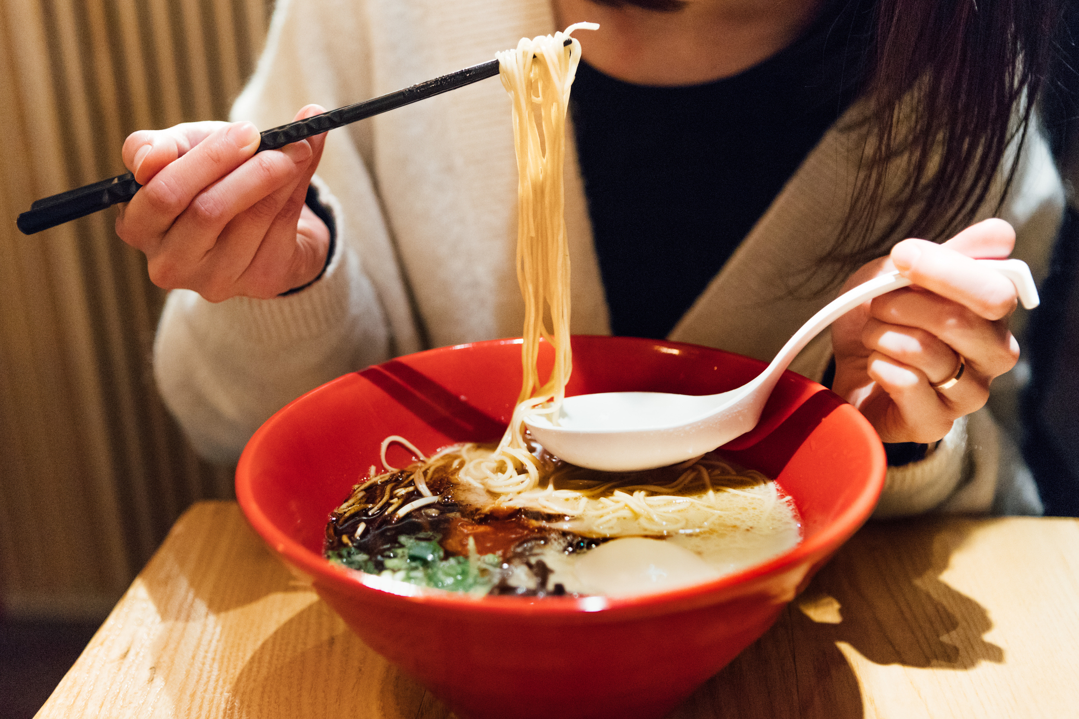 A person holding chopsticks and a spoon, eating a bowl of ramen. The focus is on the food and hands, not the person's face