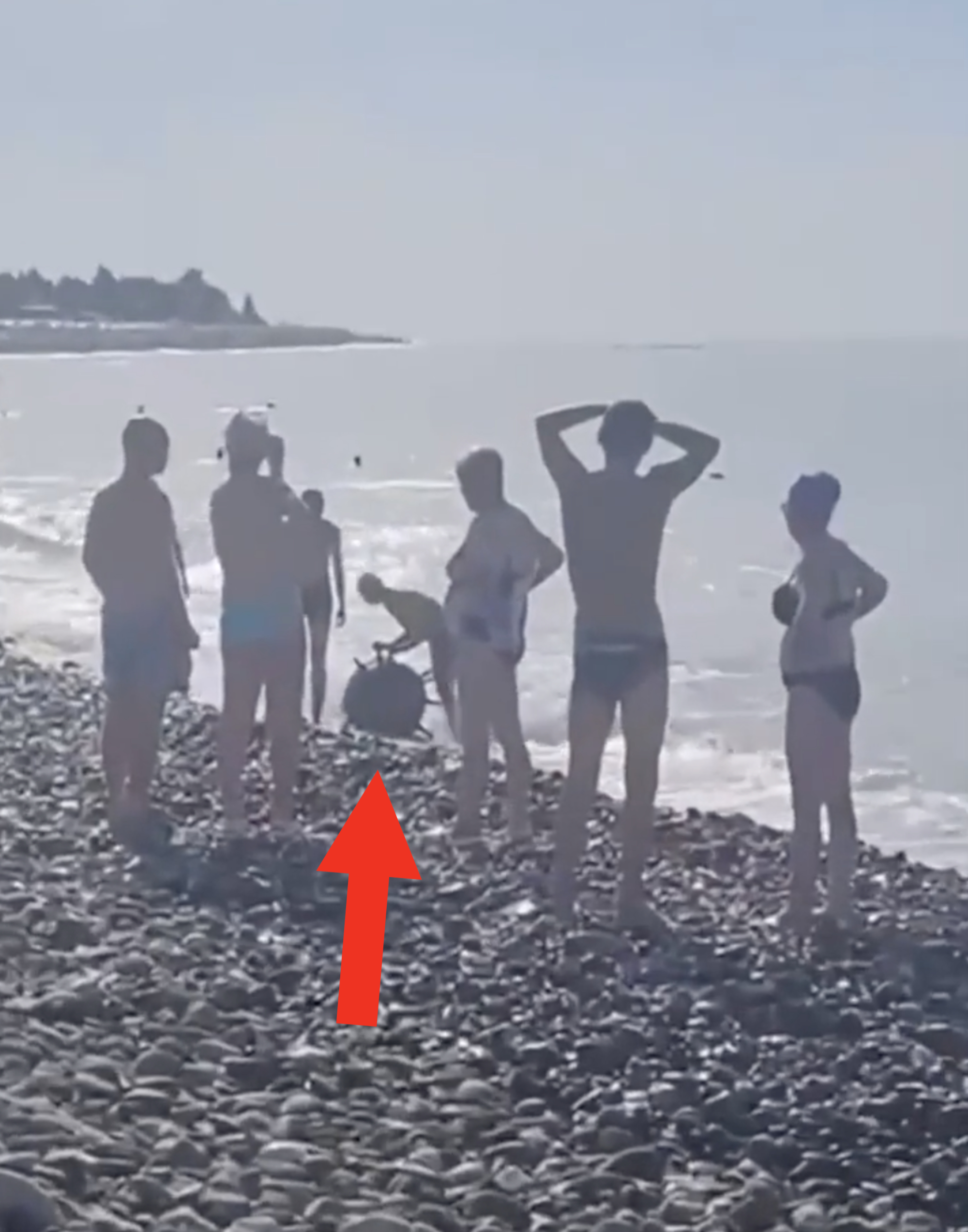 A group of people stand on a rocky beach by the water, some in swimwear, talking and relaxing