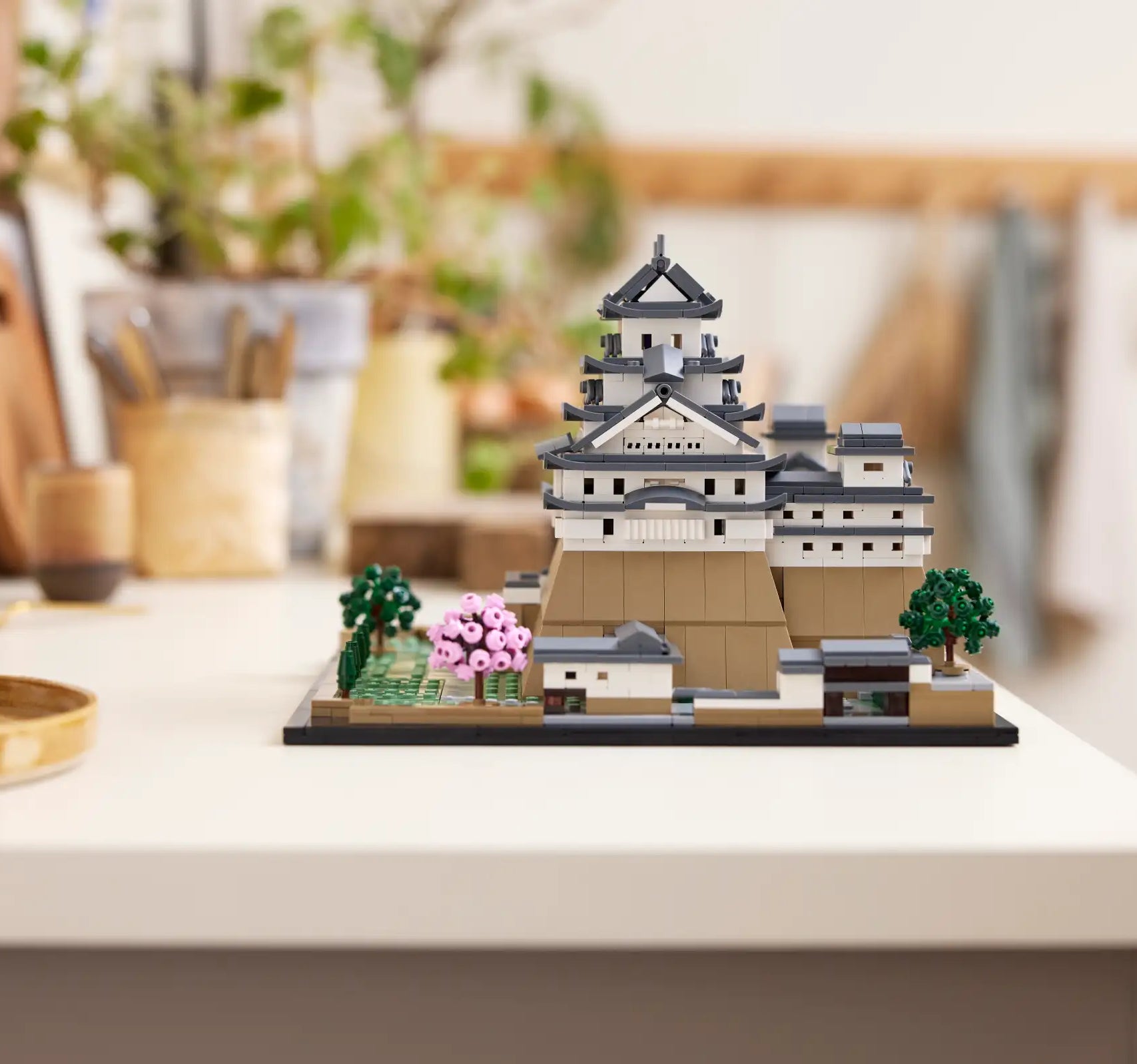 A detailed LEGO replica of a traditional Japanese castle is displayed on a white tabletop, with various household items and plants blurred in the background