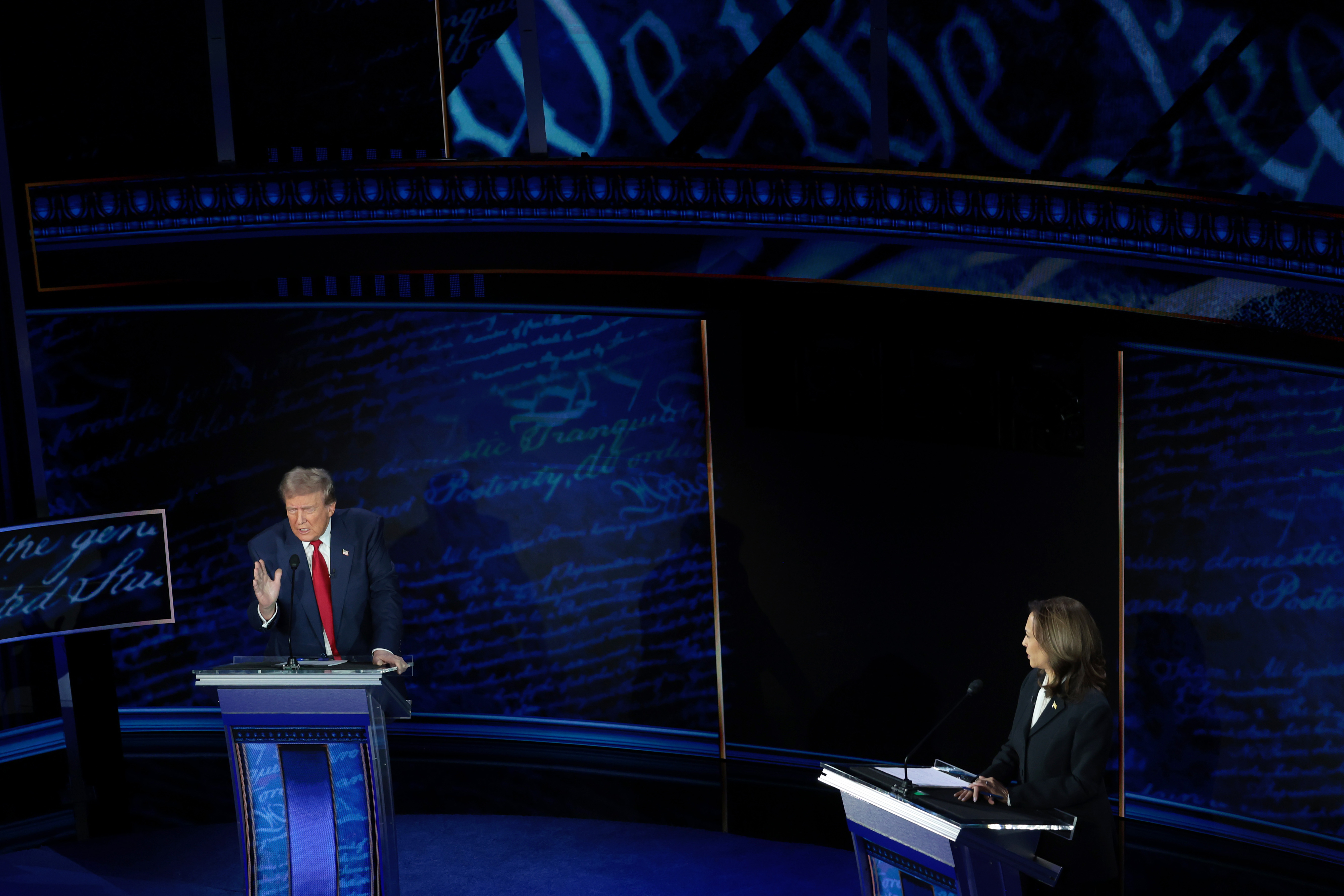Donald Trump and Kamala Harris participate in a debate, each standing at separate podiums on a stage with projected text in the background