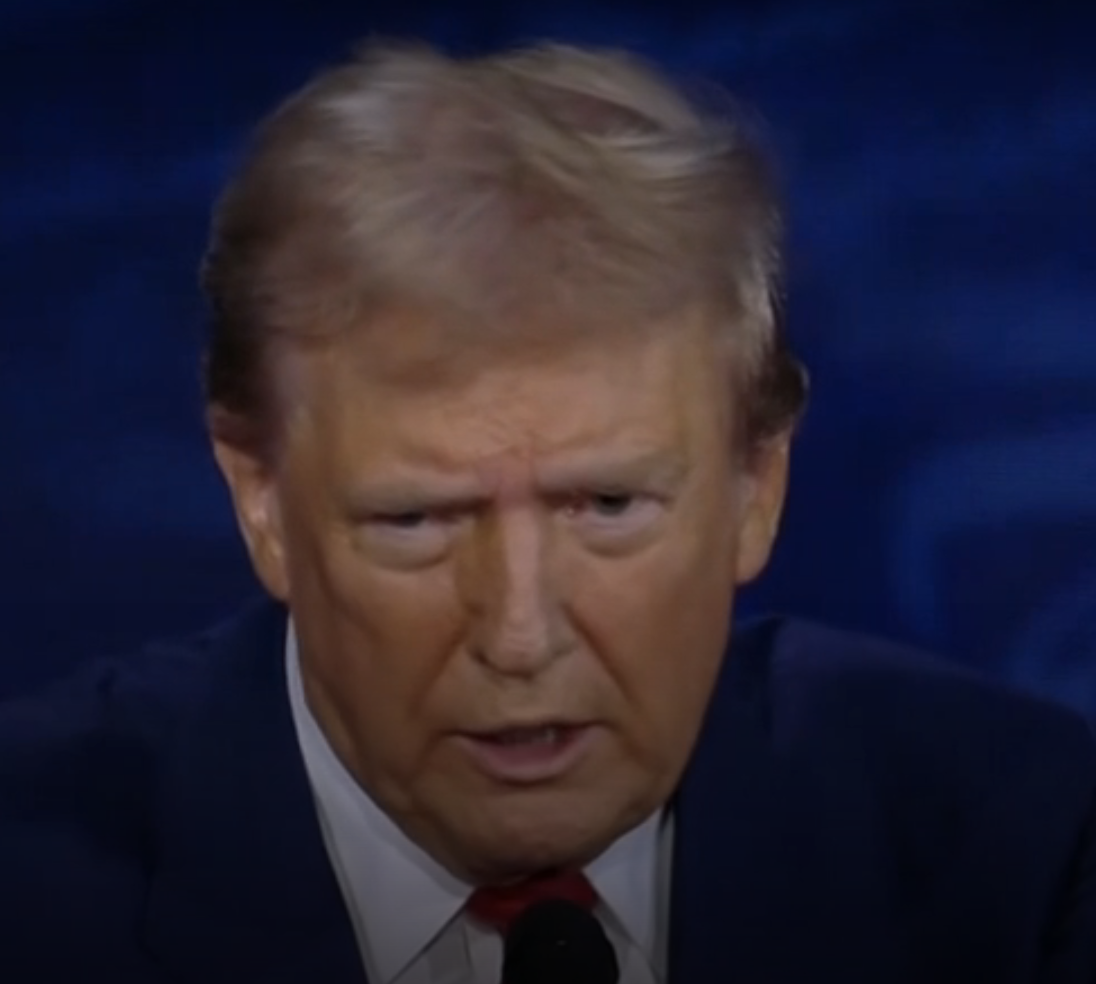 Donald Trump speaking at a public event, looking serious, wearing a dark suit and a white shirt