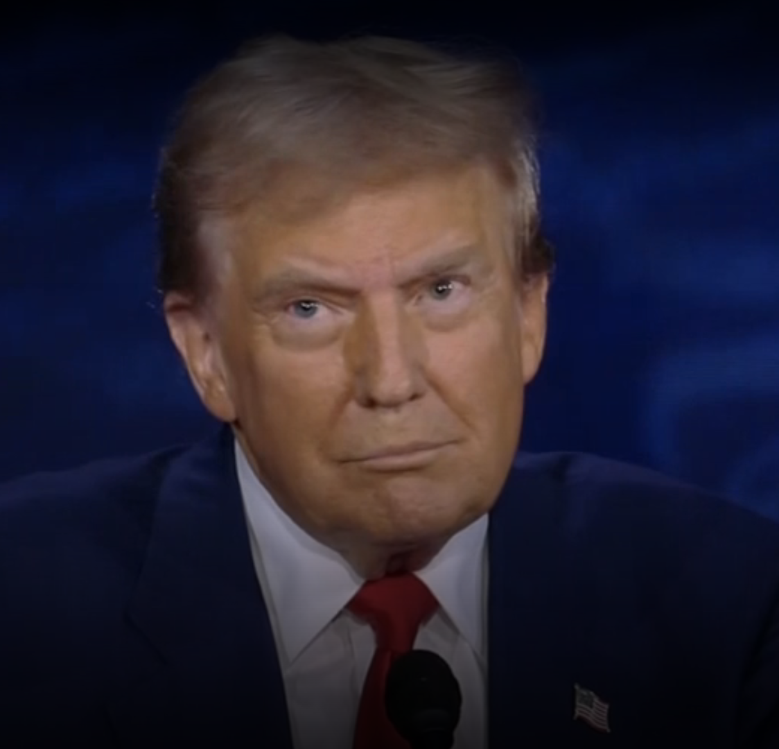 Donald Trump at a speaking event, wearing a dark suit, white shirt, and red tie, with an American flag pin on his lapel