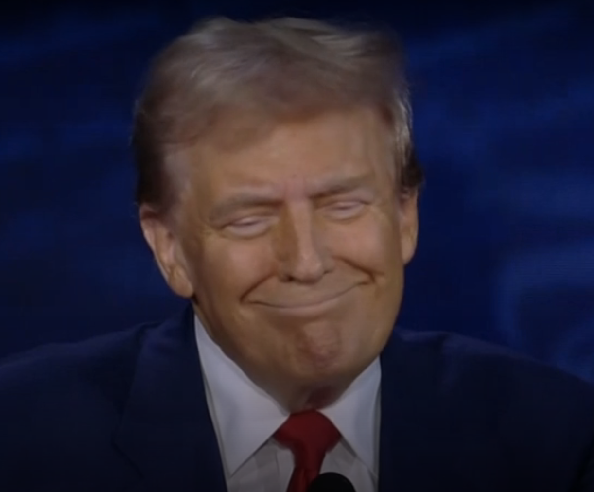 Donald Trump, in a suit and tie, is smiling with a closed mouth against a dark background