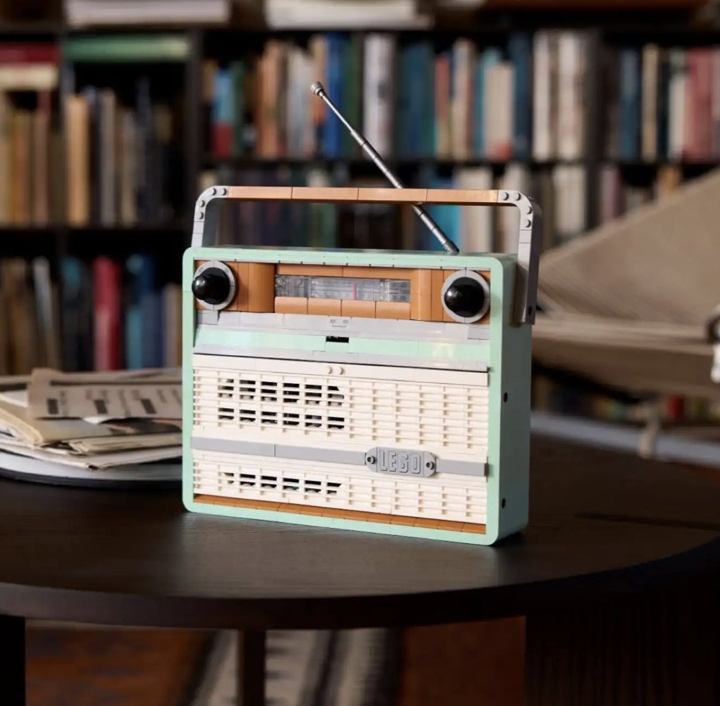 A retro-style radio sits on a round table with magazines beside it, in a cozy room lined with bookshelves and a lounge chair in the background