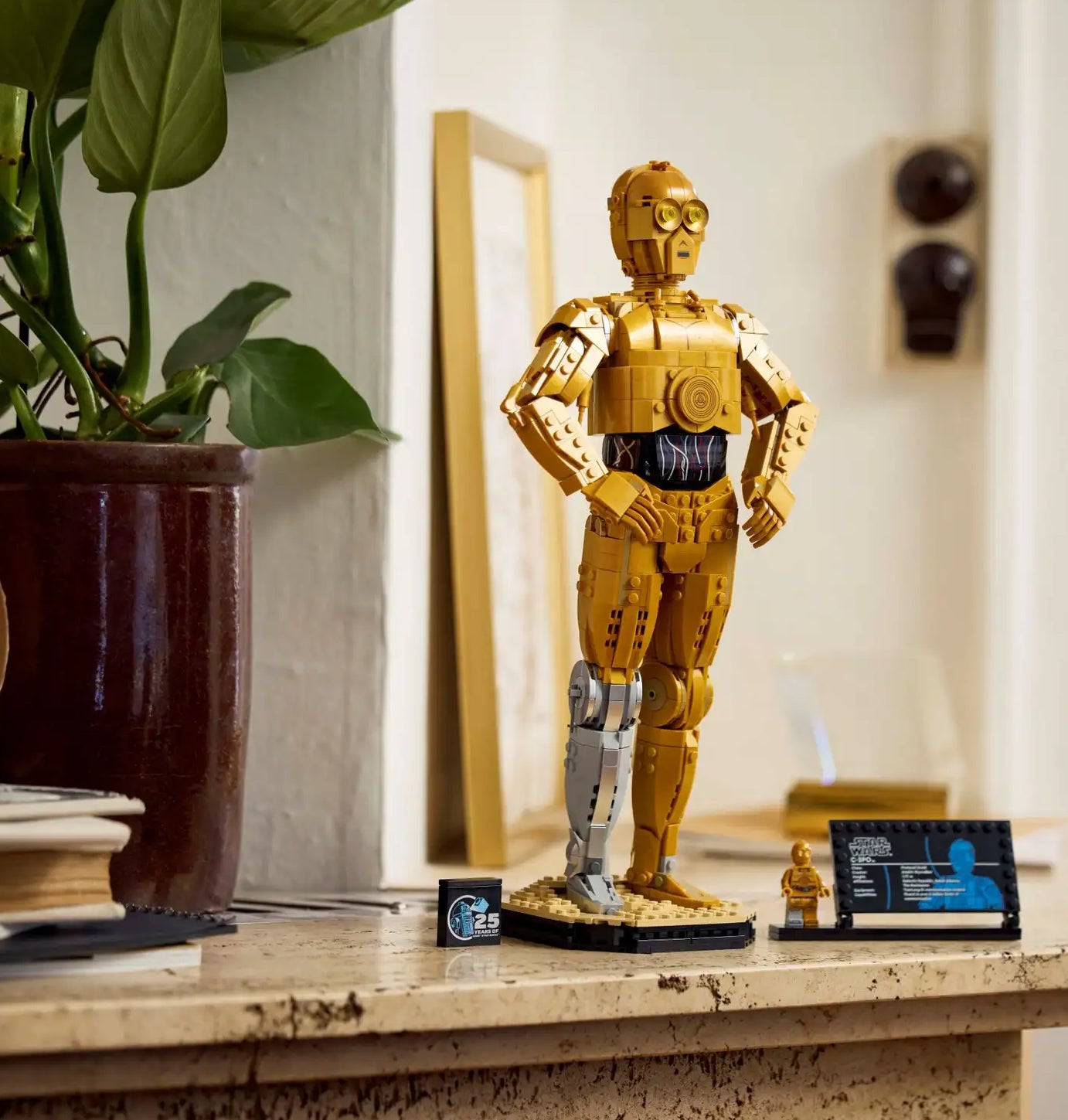Lego model of Star Wars character C-3PO is displayed on a tabletop with books, a potted plant, and a small screen in the background