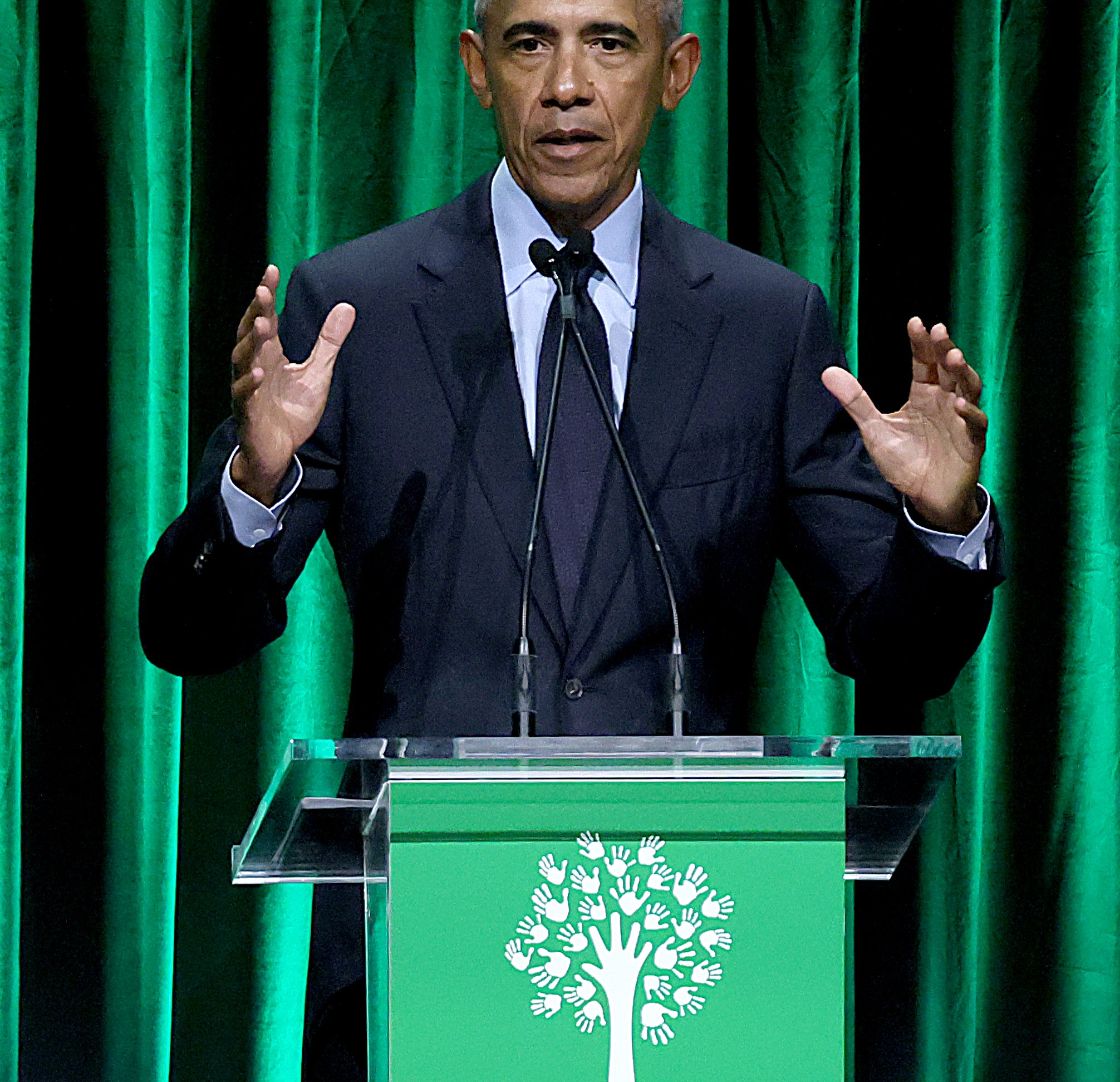 Barack Obama speaks at the Sandy Hook Promise event, standing behind a podium with a green backdrop and the Sandy Hook Promise logo