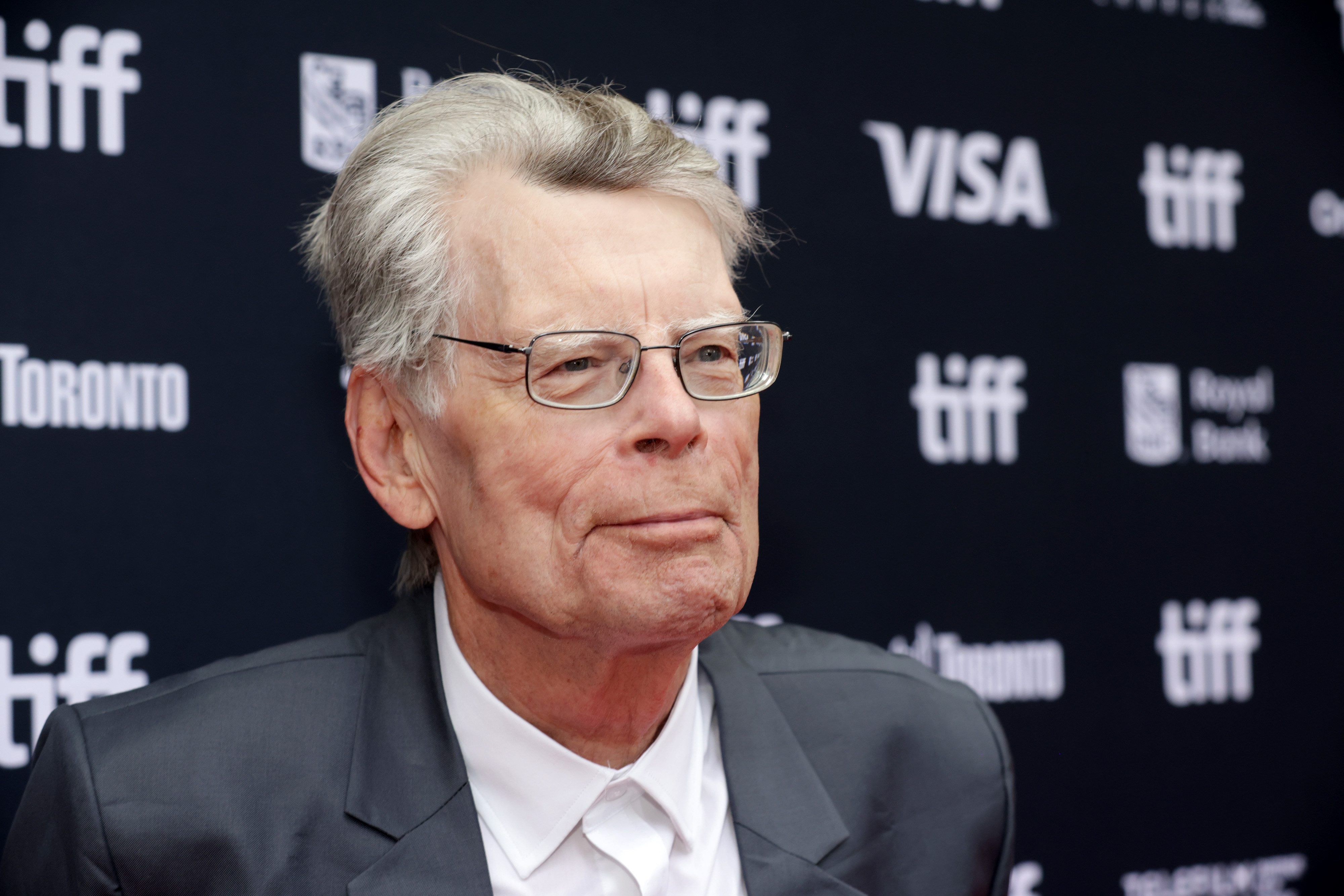 Stephen King at the Toronto International Film Festival, wearing a suit jacket and shirt, against a backdrop with TIFF and sponsor logos