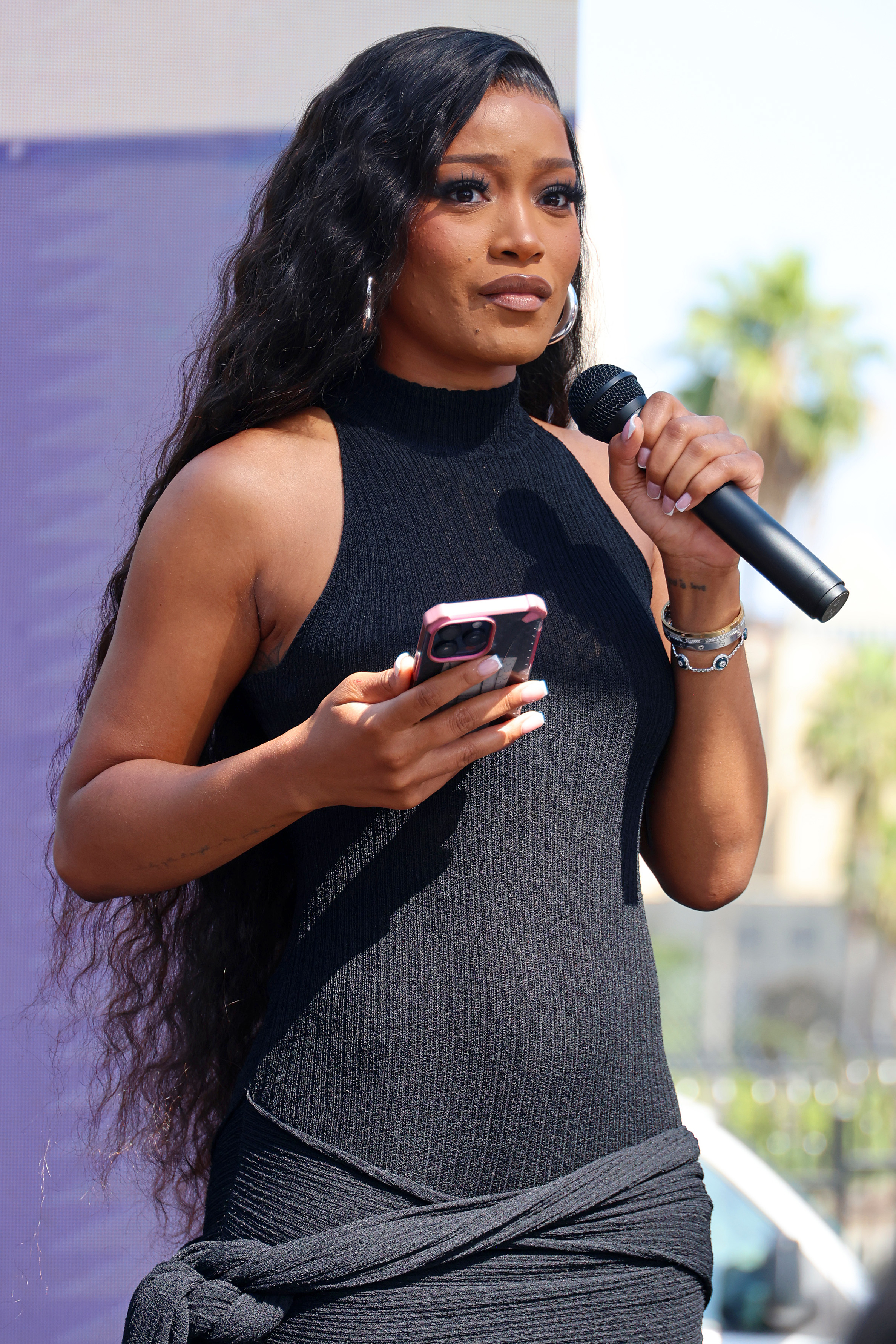 Keke Palmer speaks into a microphone while holding a smartphone. She is wearing a stylish halter dress
