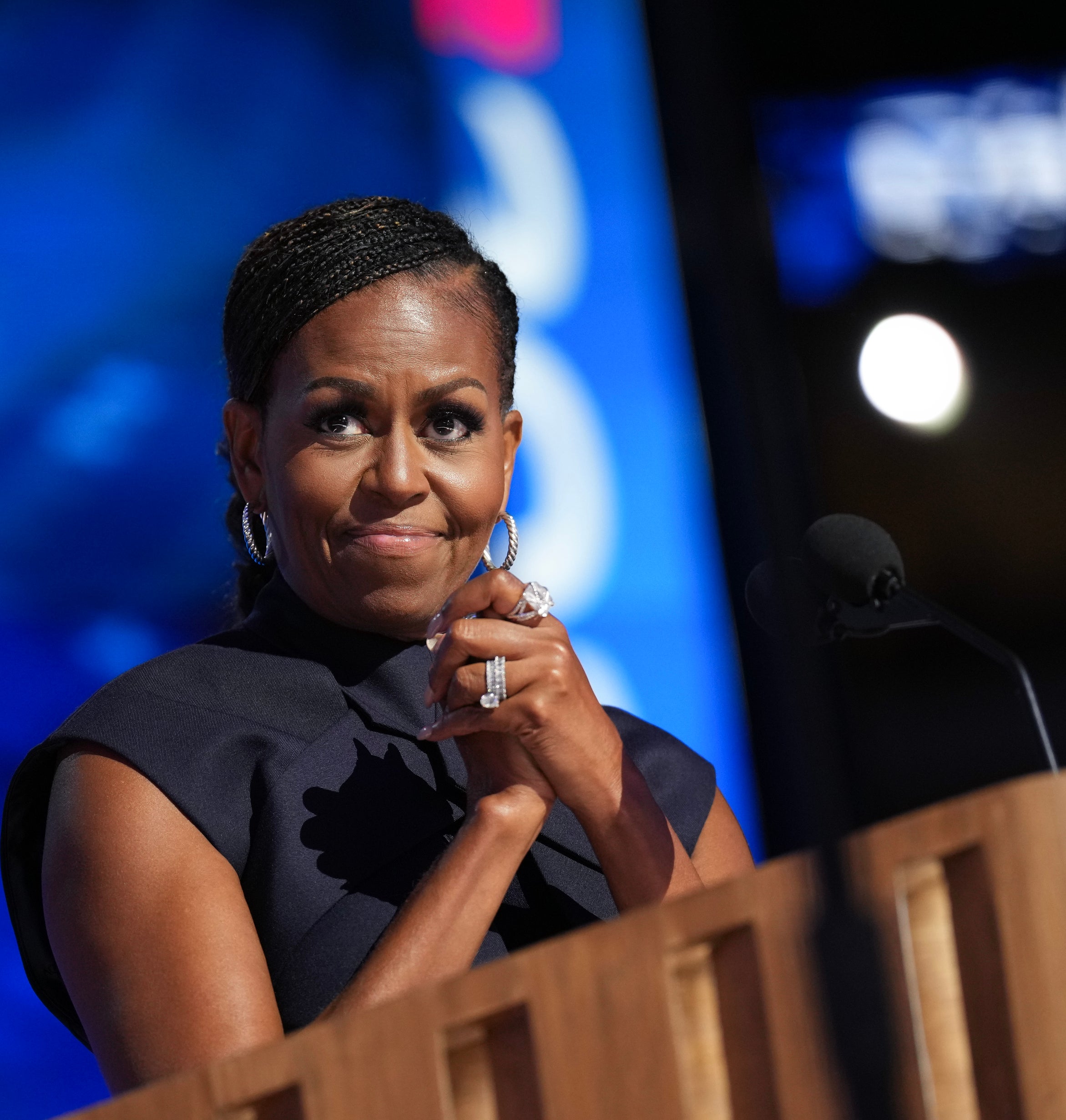 Michelle Obama smiling and clasping her hands while standing at a podium, wearing a sleeveless, high-neck dress