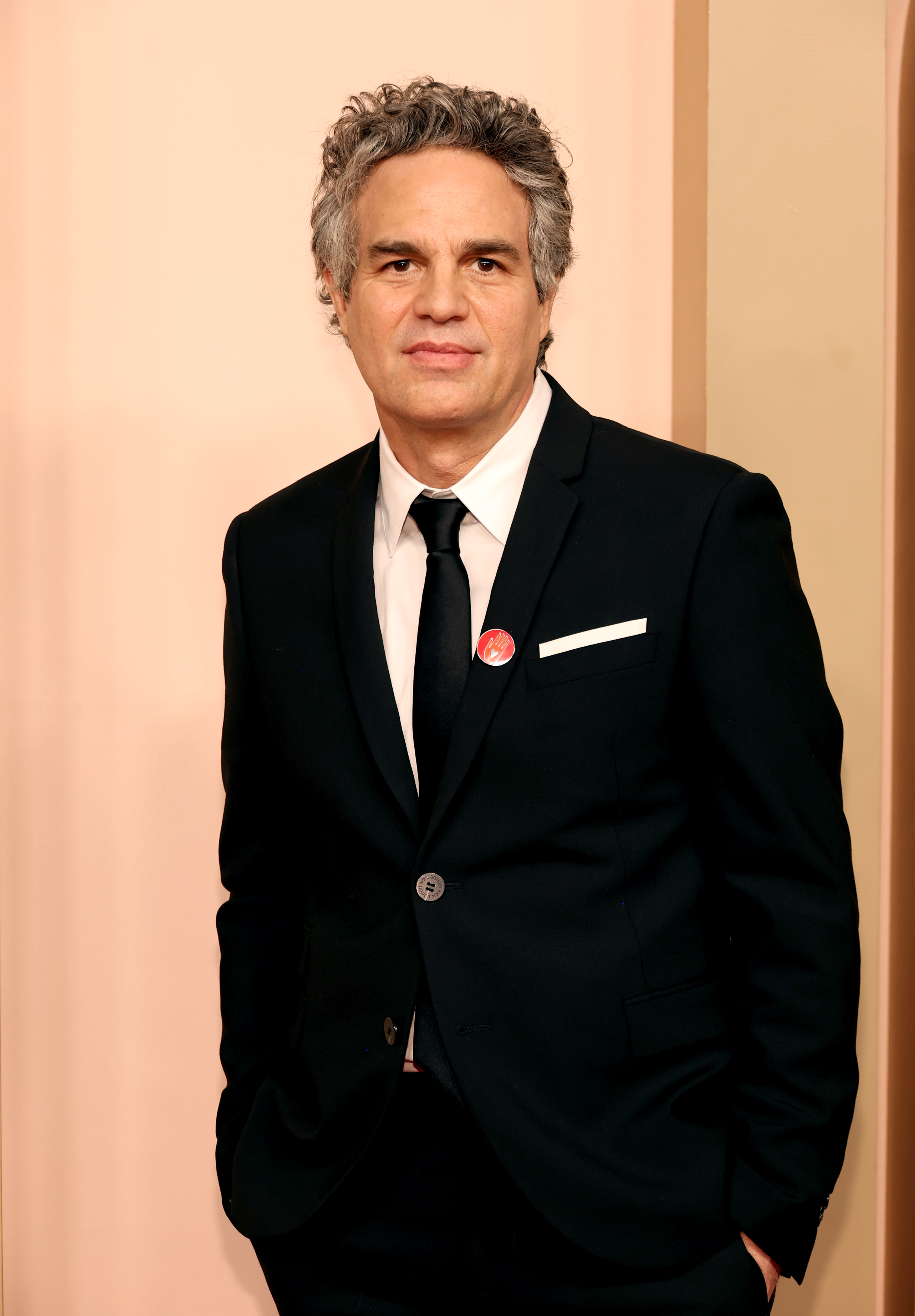 Mark Ruffalo posing on the red carpet in a dark suit with a white pocket square, black tie, and a button pin on his lapel