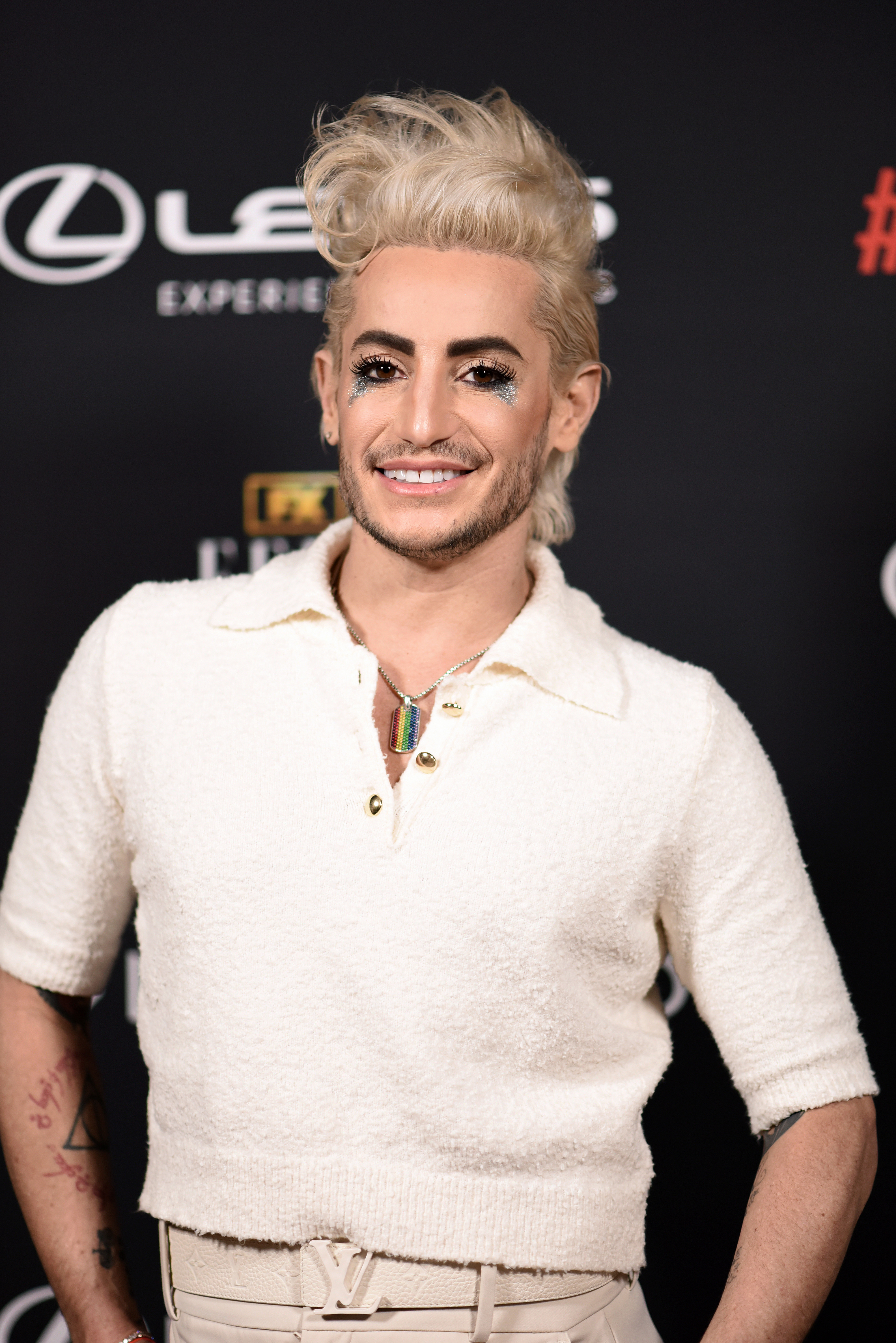 Frankie Grande on the red carpet, wearing a stylish short-sleeve top with a collar, accessorized with glittery makeup and a colorful necklace