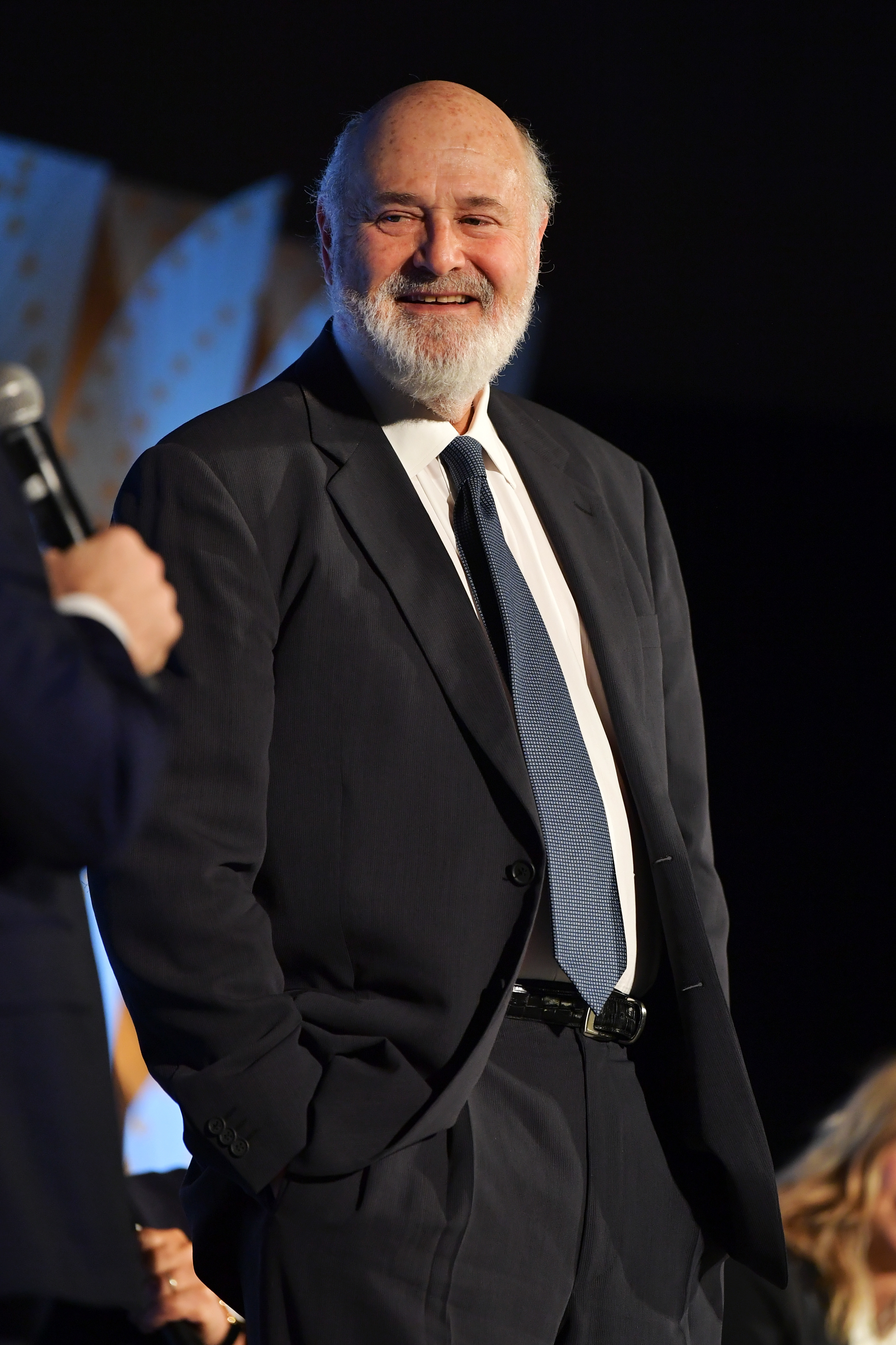 Rob Reiner smiles while wearing a suit and tie, standing with hands in pockets at an event