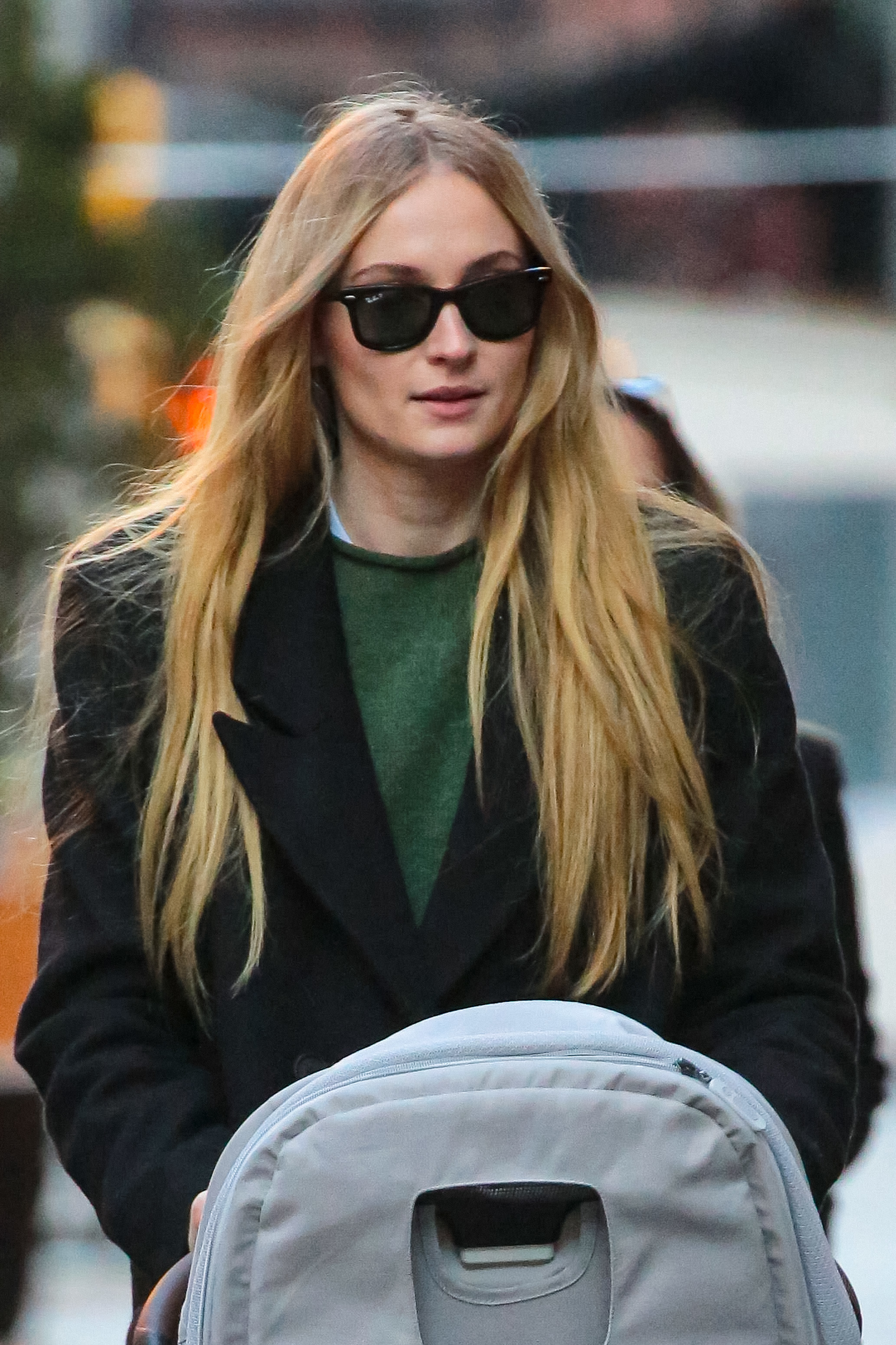 Sophie Turner is seen walking outdoors, wearing sunglasses, a long coat, and pushing a baby stroller