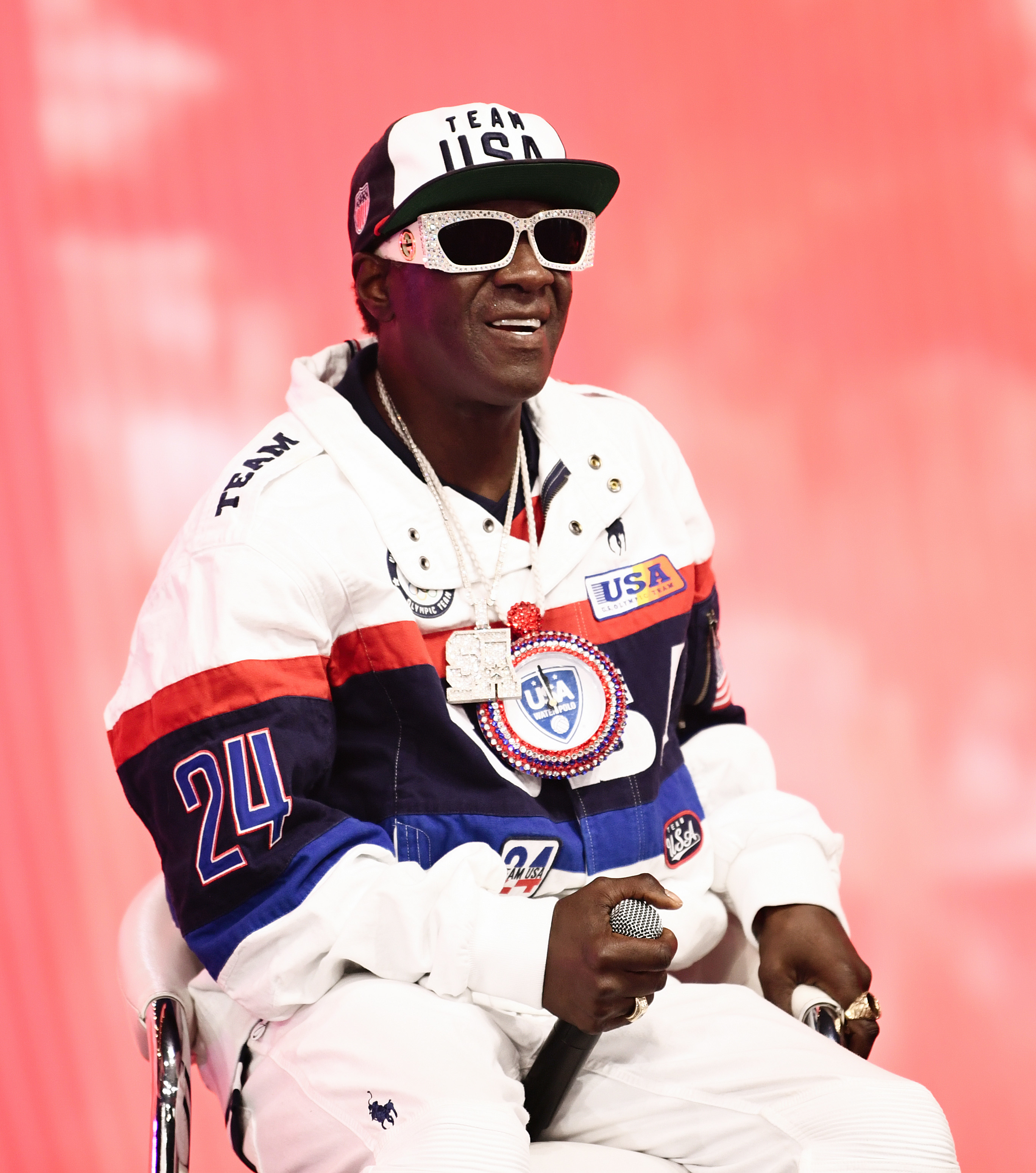 Flavor Flav wears a white jacket with a &quot;24&quot; on the sleeve, &quot;Team USA&quot; cap, and large sunglasses, holding a mic