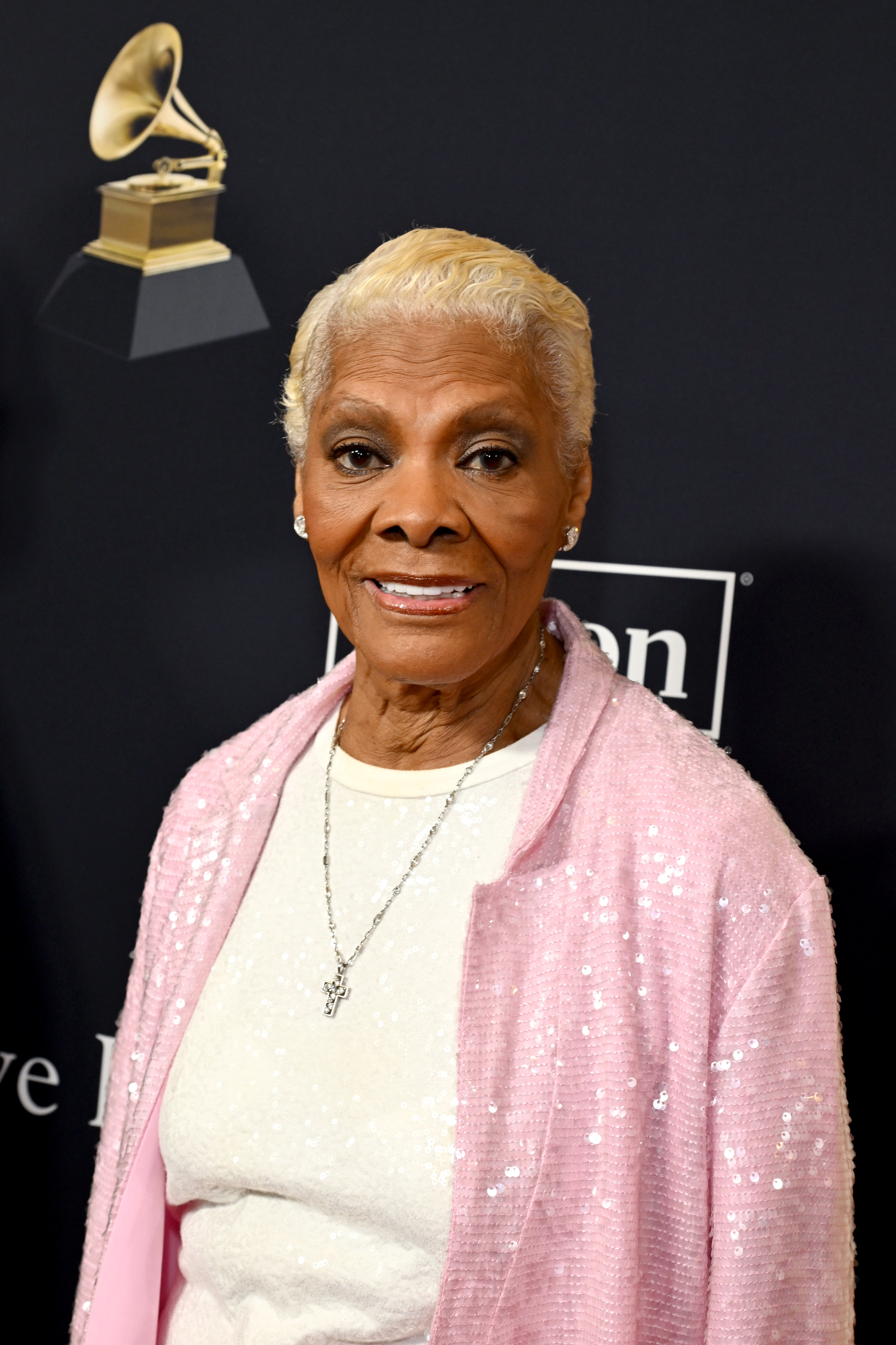 Dionne Warwick at a Grammy event, wearing a sparkling jacket over a top, smiling for the camera. The Grammy logo is visible in the background