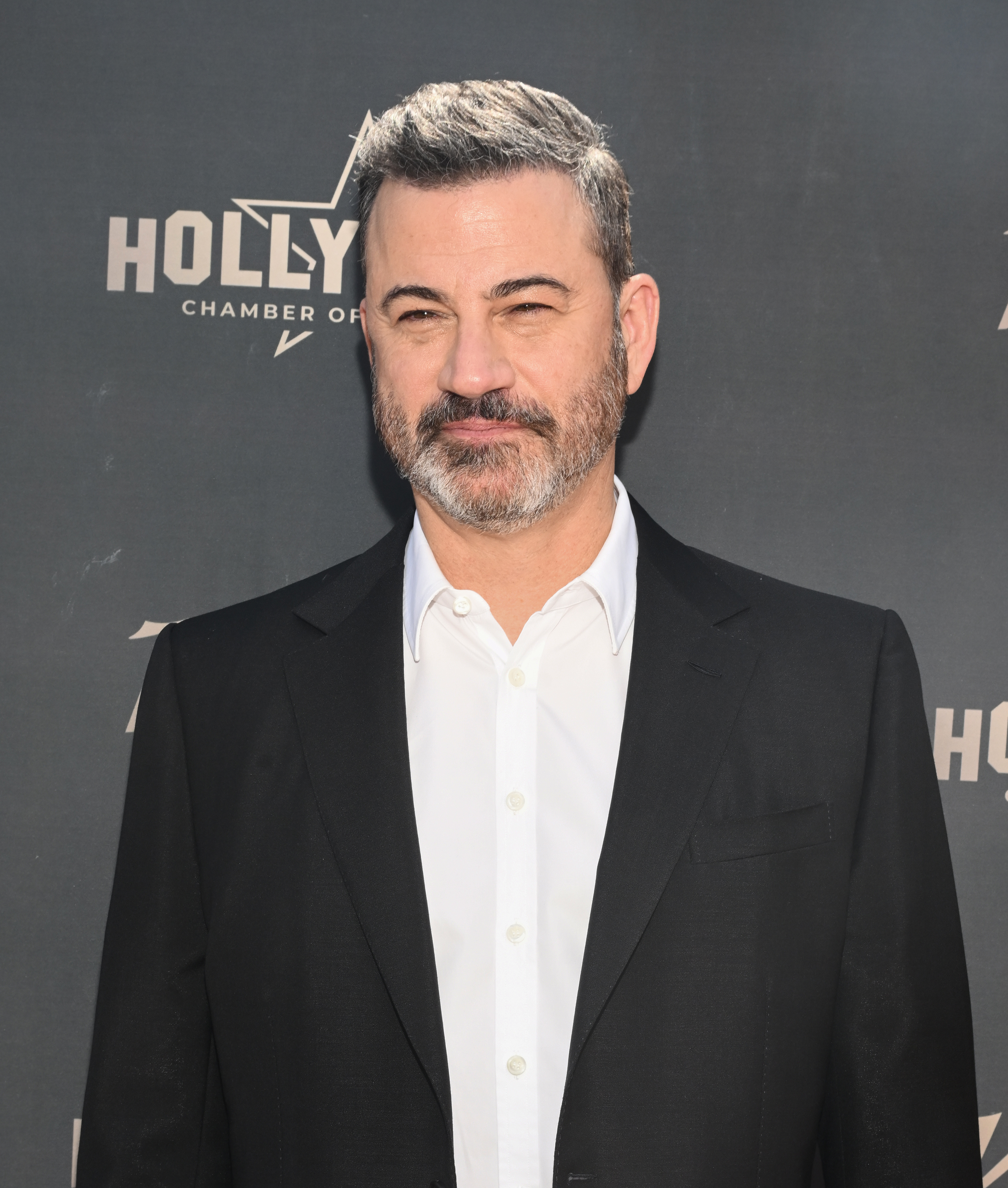 Jimmy Kimmel on a red carpet, wearing a dress shirt and a black suit jacket, with a Hollywood Chamber of Commerce backdrop