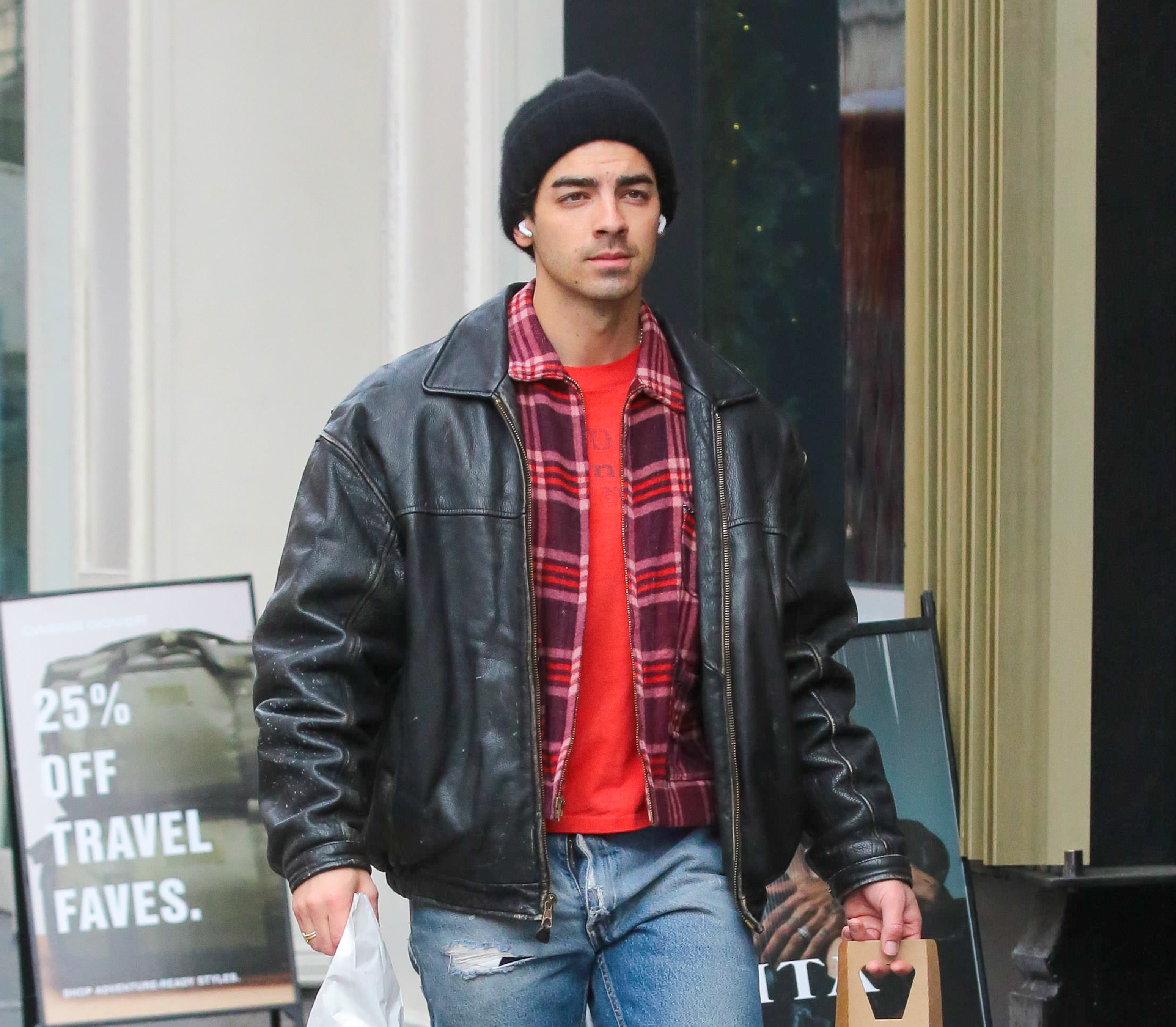 Joe Jonas wearing a dark leather jacket and ripped jeans, holding a shopping bag while walking outside