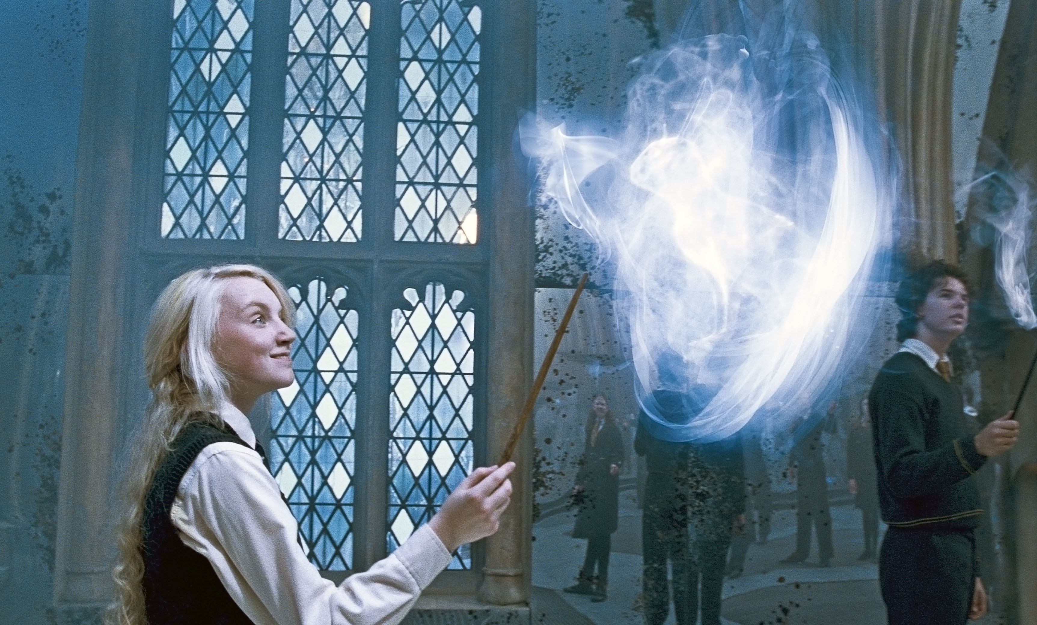 Evanna Lynch as Luna Lovegood conjures a Patronus charm with her wand in a large, ornate room with tall windows. Other students practice spells in the background
