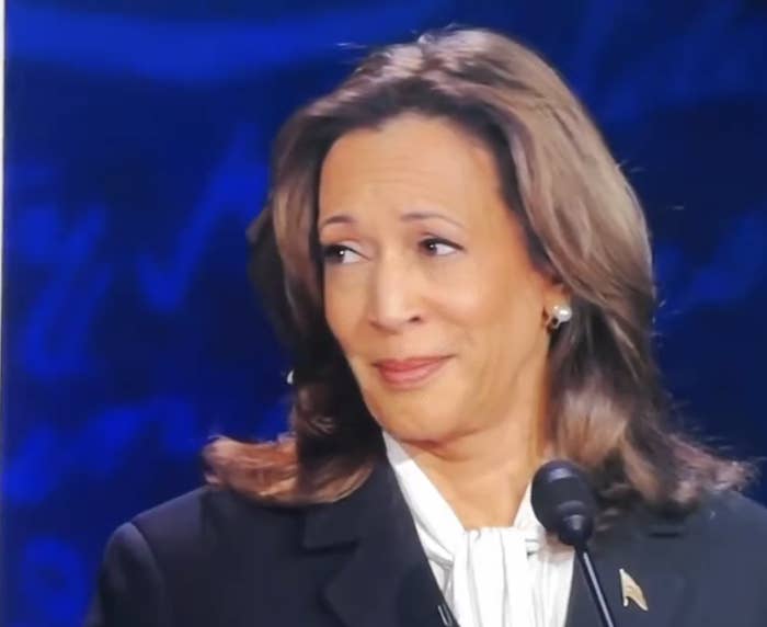 Kamala Harris, wearing a white blouse and dark blazer, stands in front of a microphone with a neutral expression against a blurred blue background