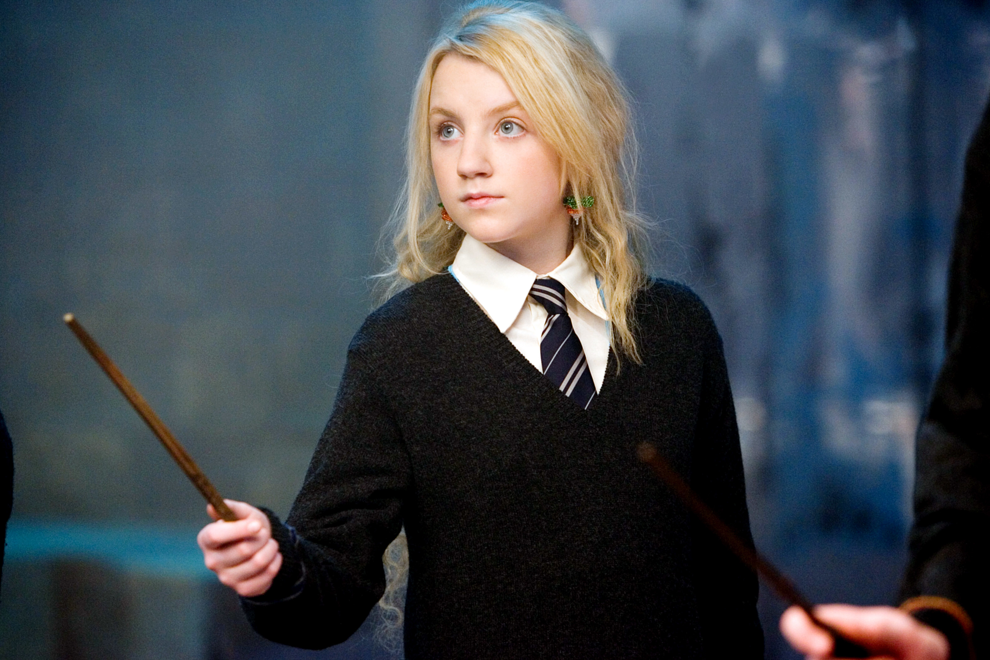 Evanna Lynch holding a wand, dressed as Luna Lovegood in a school uniform with a tie, looking attentive