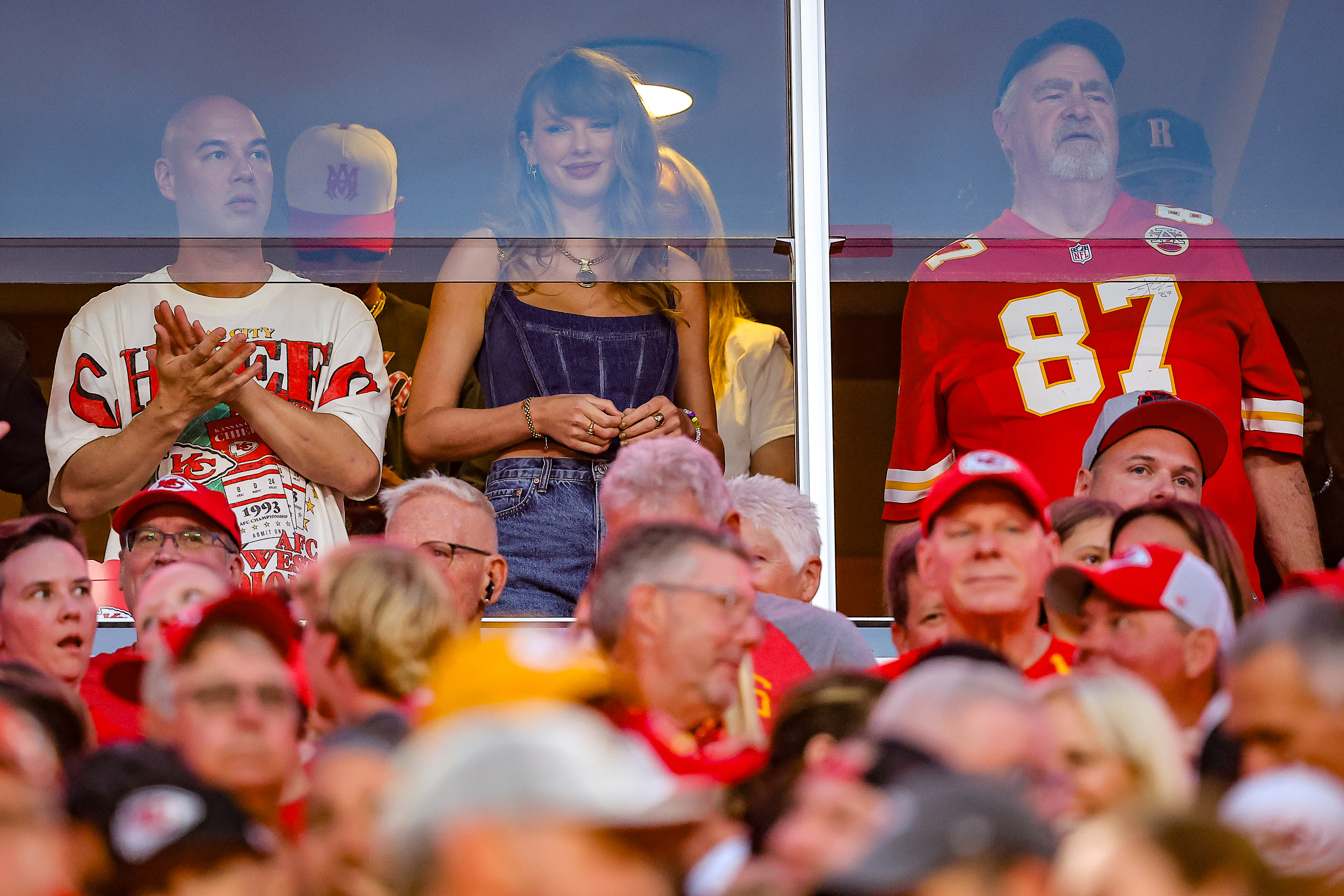 Taylor Swift in a suite at a football game, wearing a stylish casual outfit, standing between two men in Kansas City Chiefs jerseys