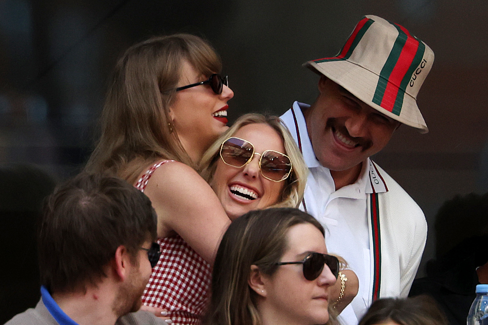 Taylor Swift, Blake Lively, and Hugh Jackman smiling and hugging at a public event. Swift wears a checkered dress, Lively wears sunglasses, and Jackman wears a Gucci hat
