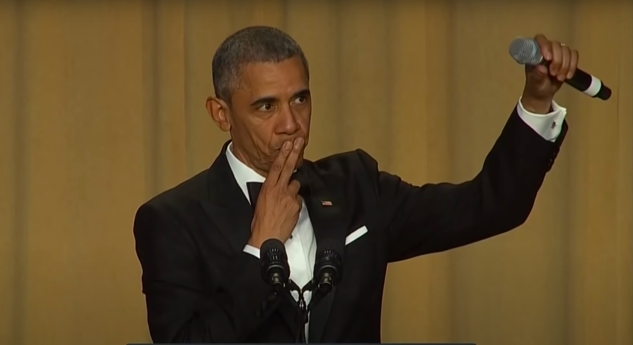 Barack Obama in a tuxedo, standing at a podium, raising a microphone, and making a gesture with his other hand