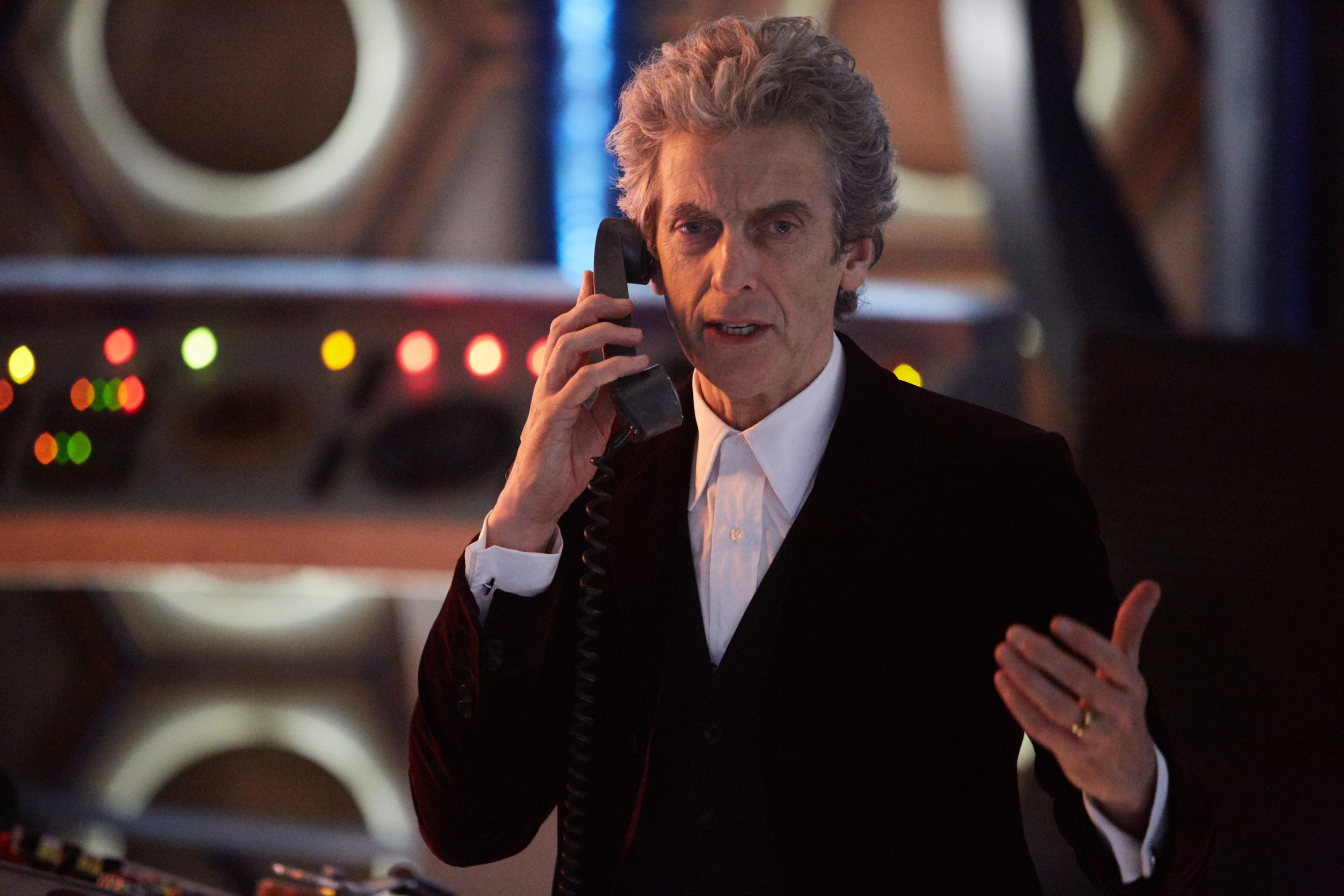 Peter Capaldi in a formal suit holding a phone to his ear, set against a futuristic backdrop with flashing lights