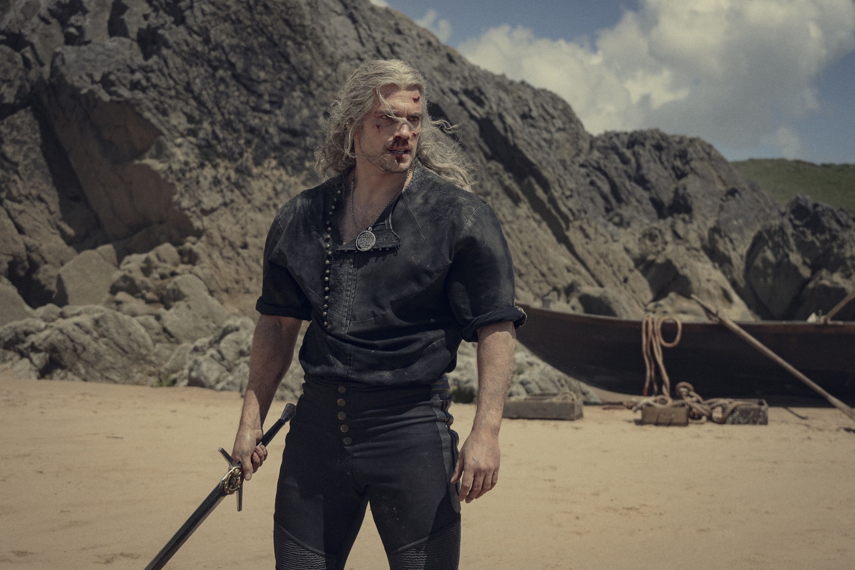 Henry Cavill as Geralt of Rivia, wearing dark casual medieval-style clothing and holding a sword, stands on a beach with rocky cliffs in the background