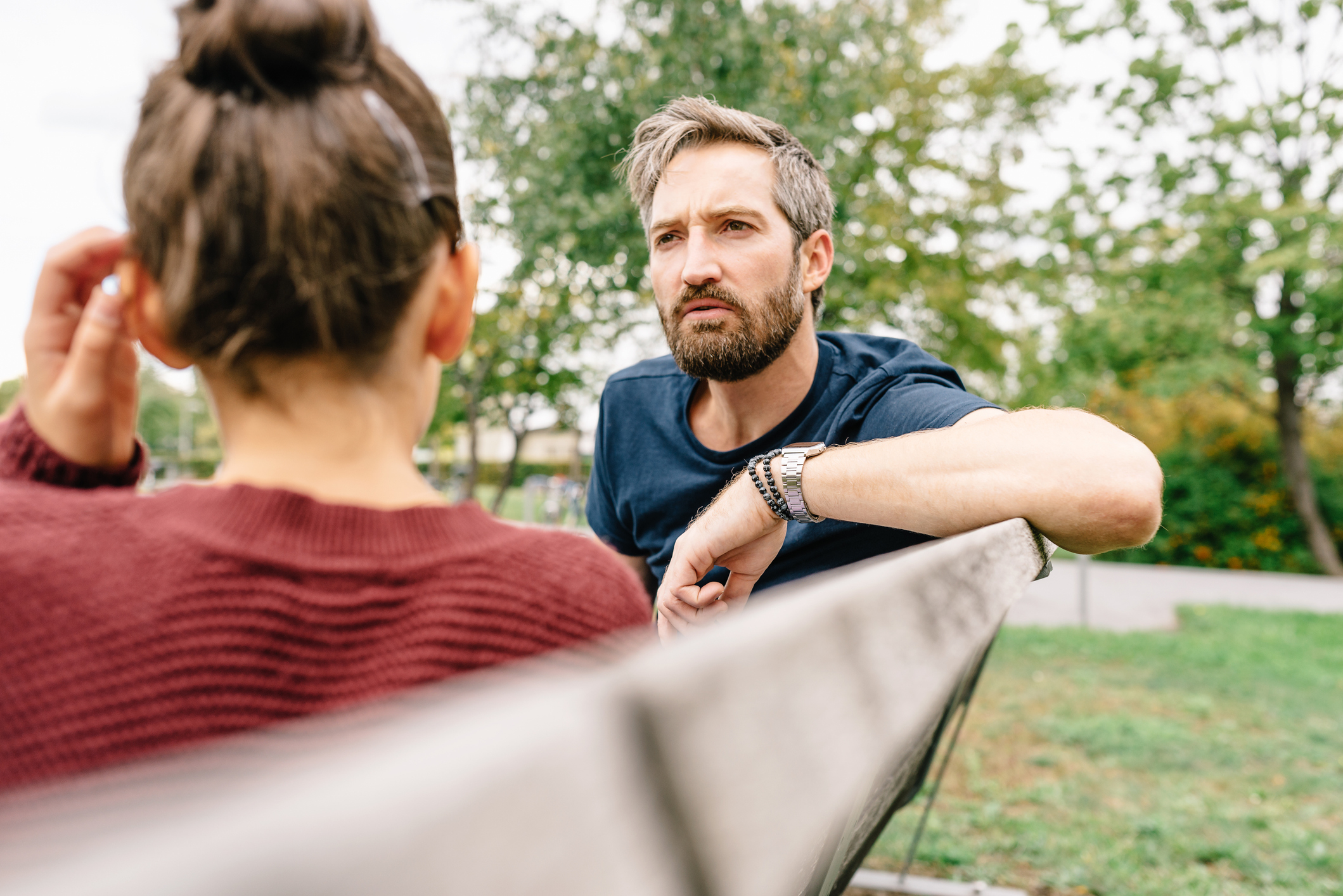 Man with beard in casual wear sits on a bench, facing a woman in glasses and sweater, engaged in serious conversation at a park