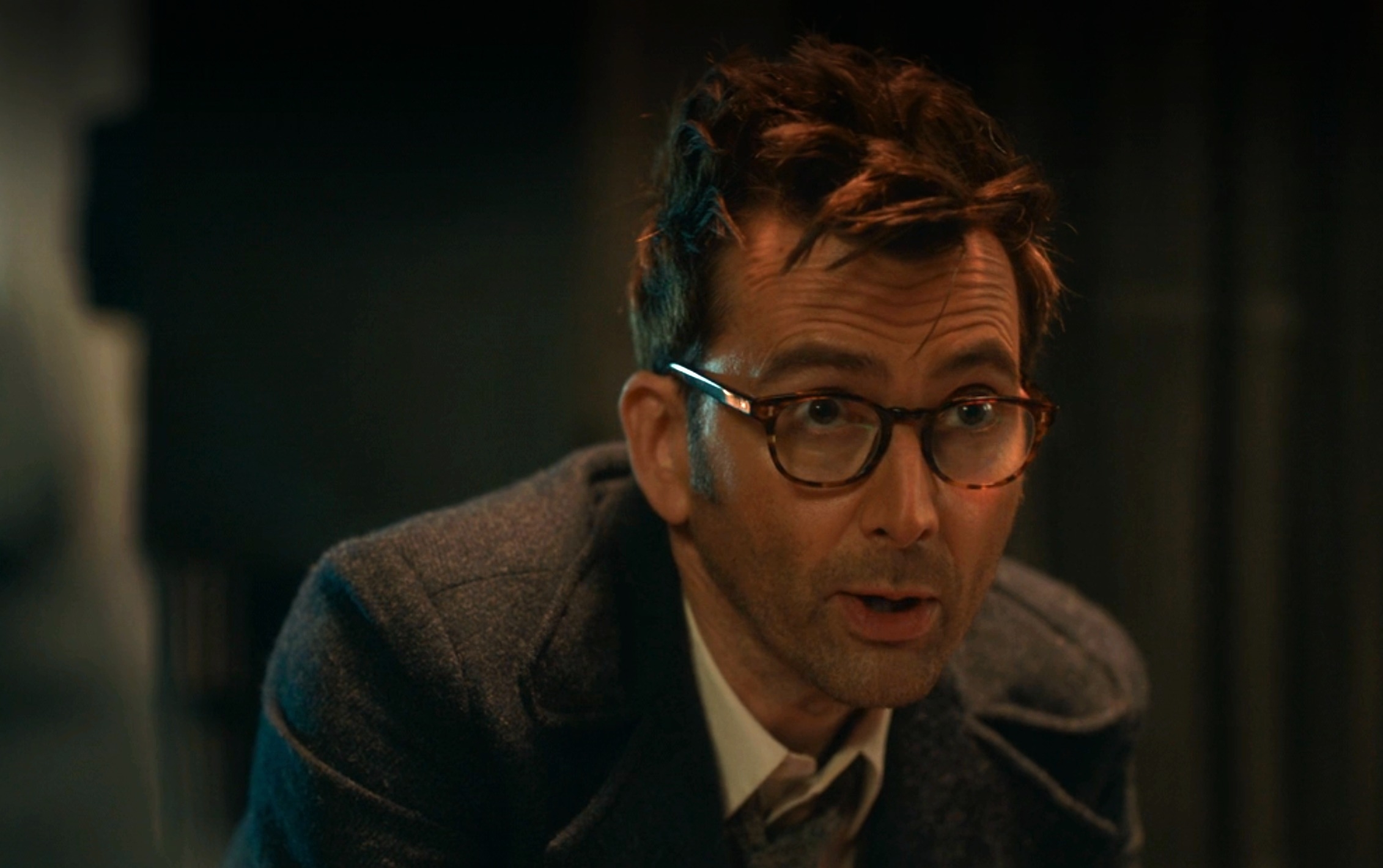 David Tennant wearing glasses and a suit, looking slightly to the side with a thoughtful expression