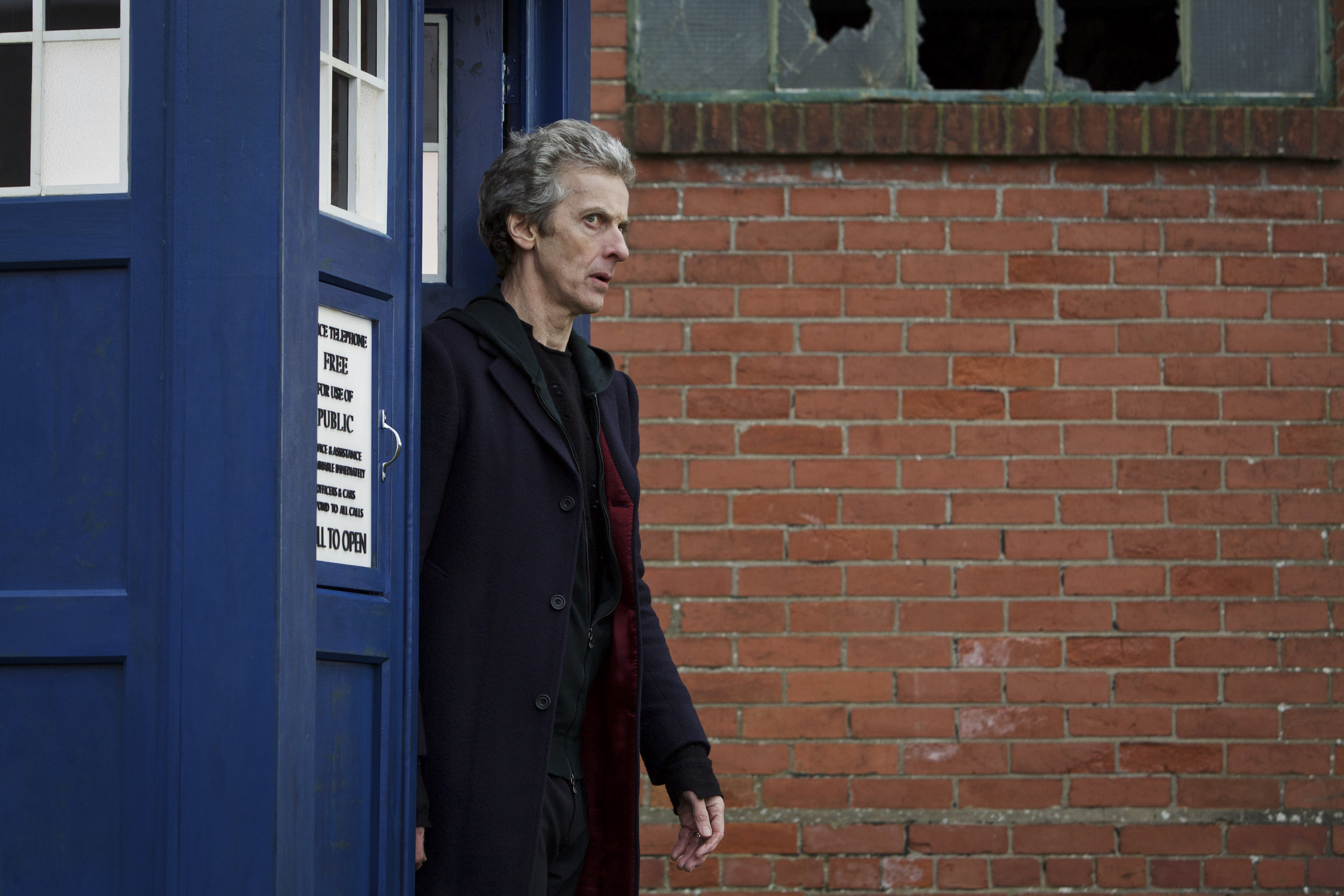 Peter Capaldi as the Doctor exits the TARDIS, a police box, with a brick wall in the background