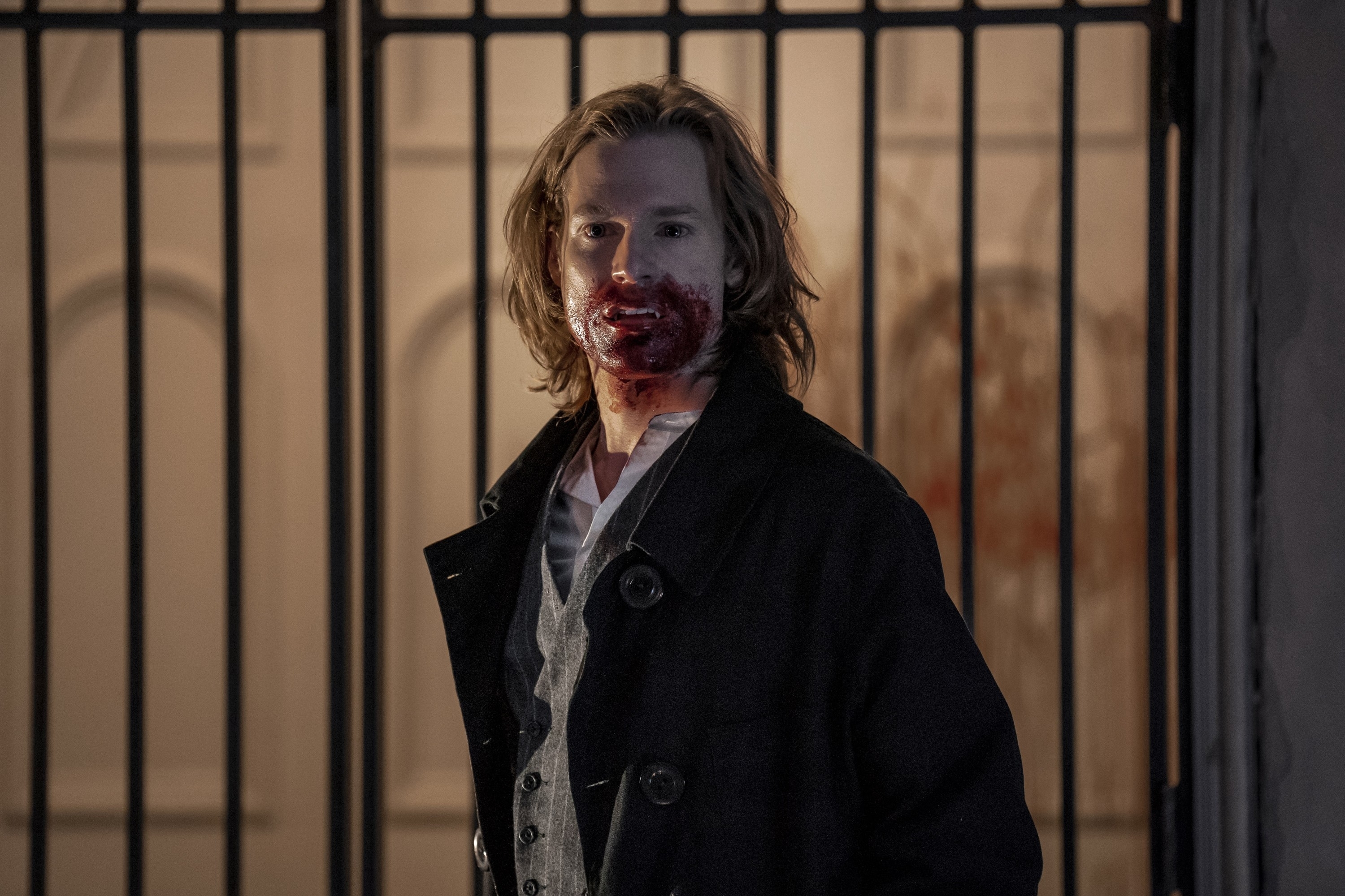 Sam Reid stands in front of a gate, wearing a dark coat over a light-colored shirt, with blood smeared on his mouth