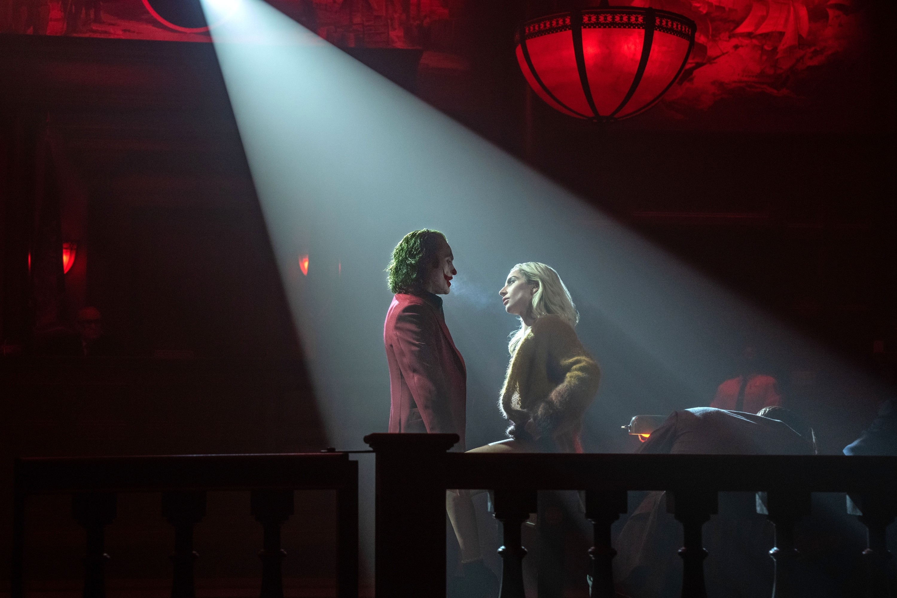 Joaquin Phoenix, in a suit, and Lady Gaga, in a fur coat, stand under a spotlight in a dramatic performance scene