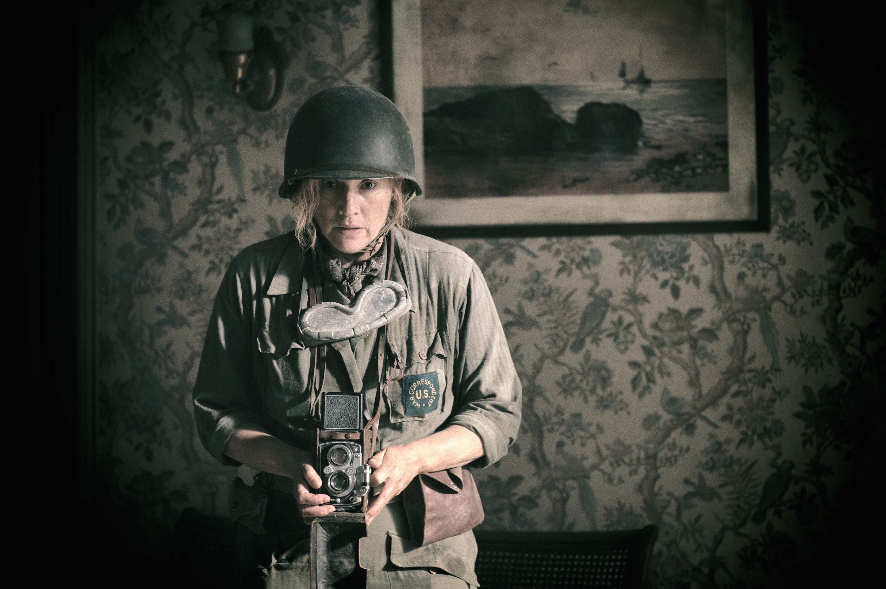 A person dressed in military gear, wearing a helmet and holding a vintage camera, stands in a room with floral wallpaper and a seaside painting in the background