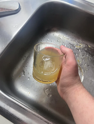 A hand holding a dirty drinking glass above a kitchen sink with water droplets and food residue