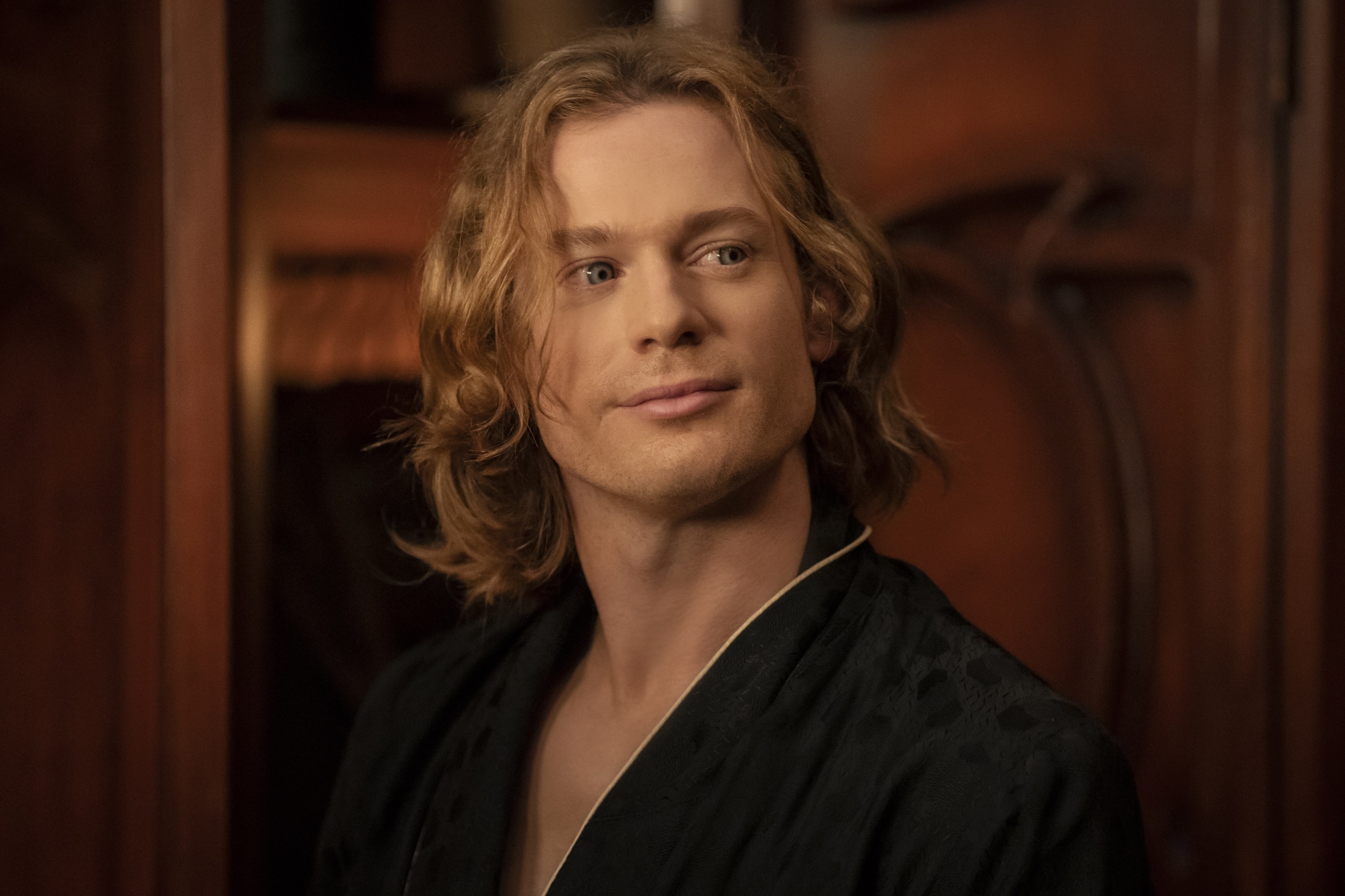 Sam with shoulder-length hair wearing a dark robe looks to the side with a slight smile. The setting appears to be indoors with wooden furniture in the background