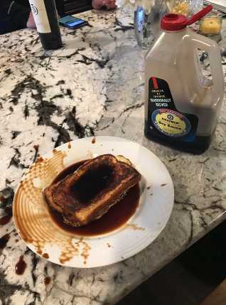 A plate with French toast soaked in syrup next to a large spilled bottle of syrup on a marble countertop. A phone and flowers are in the background