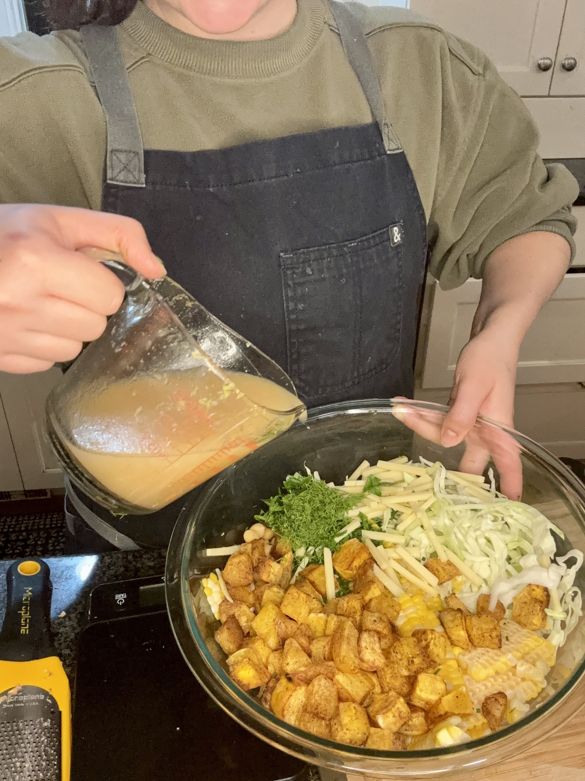 Person in a green top and black apron pours dressing into a bowl of chopped vegetables and roasted potatoes