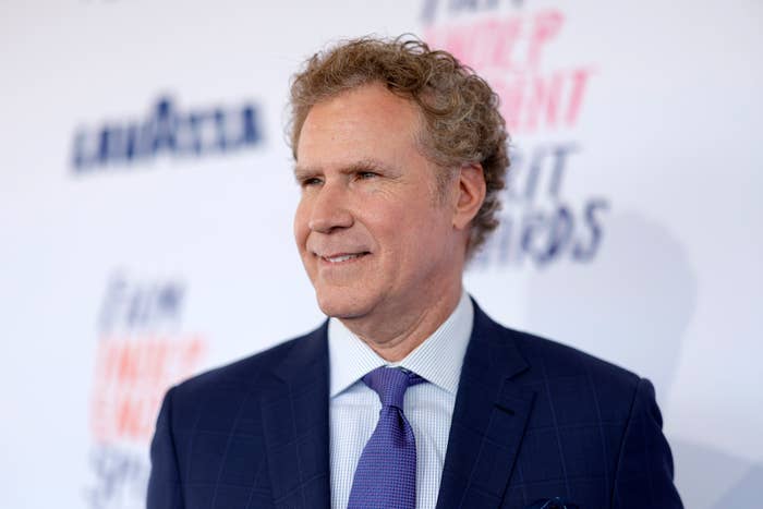 Will Ferrell in a suit and tie at a red carpet event, smiling with a neutral background displaying indistinct text