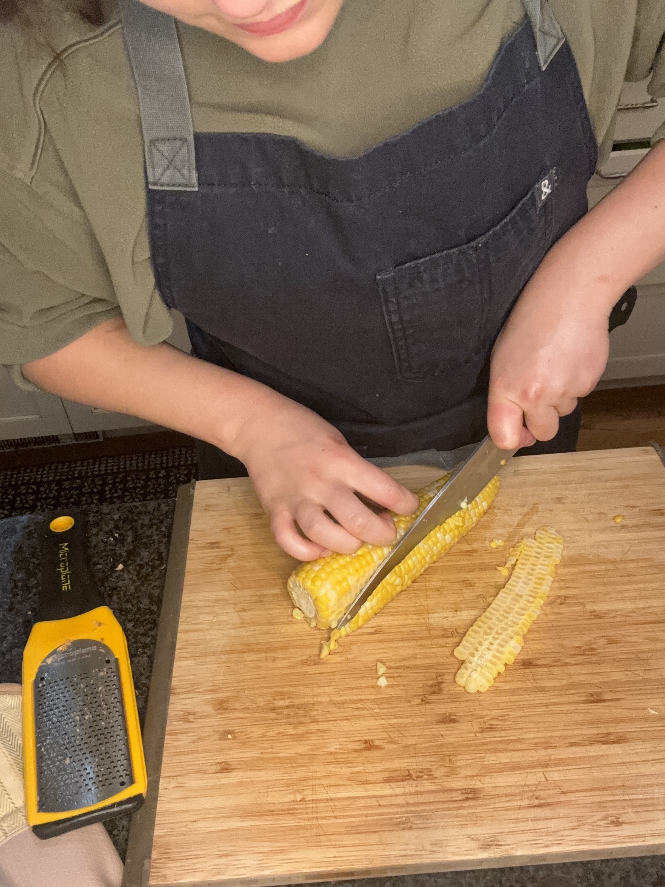 Person cutting corn off the cob on a wooden cutting board in a kitchen, with a grater and a cloth nearby
